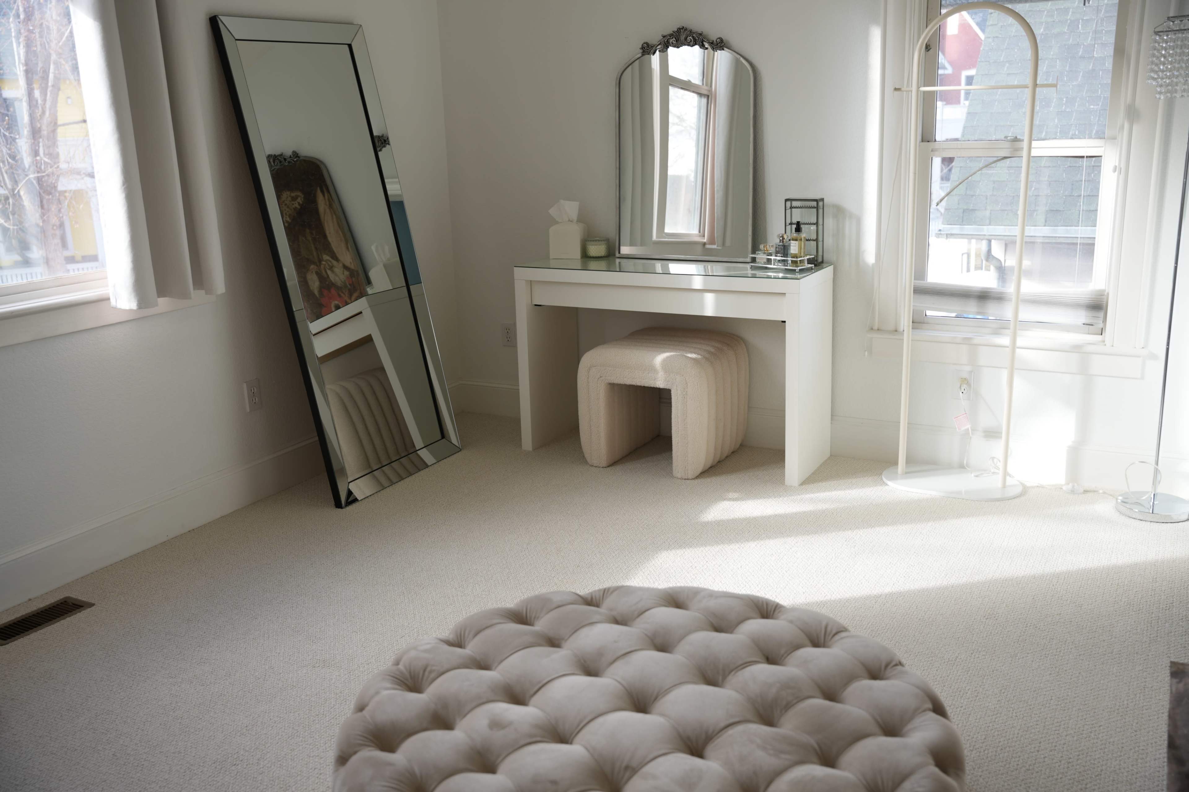A bright, minimalistic room featuring a white dressing table with a mirror, a reflective floor mirror, and a tufted ottoman.