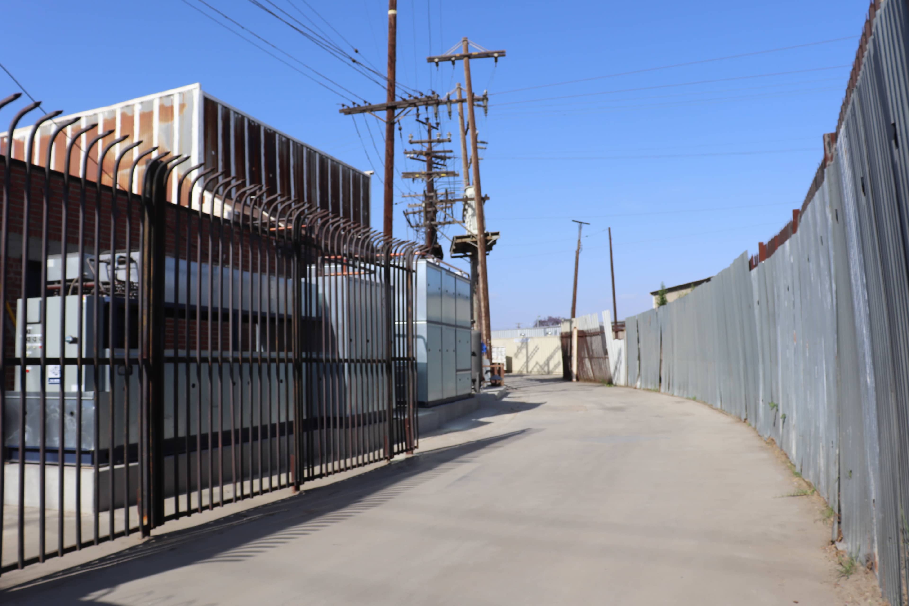 A deserted industrial alley lined with tall fences and power lines under a clear blue sky.