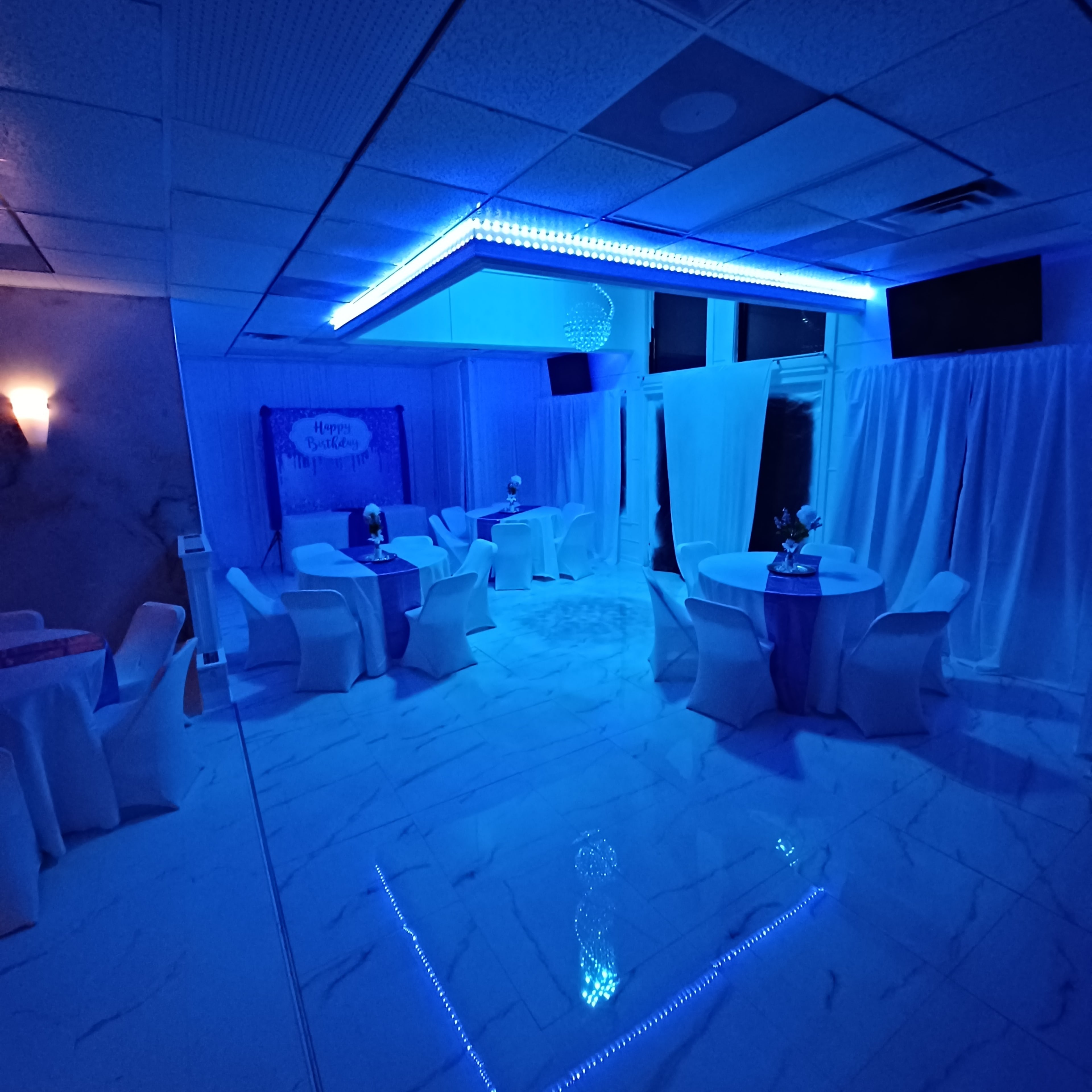 The interior of a softly lit event space features several round tables with white tablecloths and chair covers, illuminated by blue LED lighting.