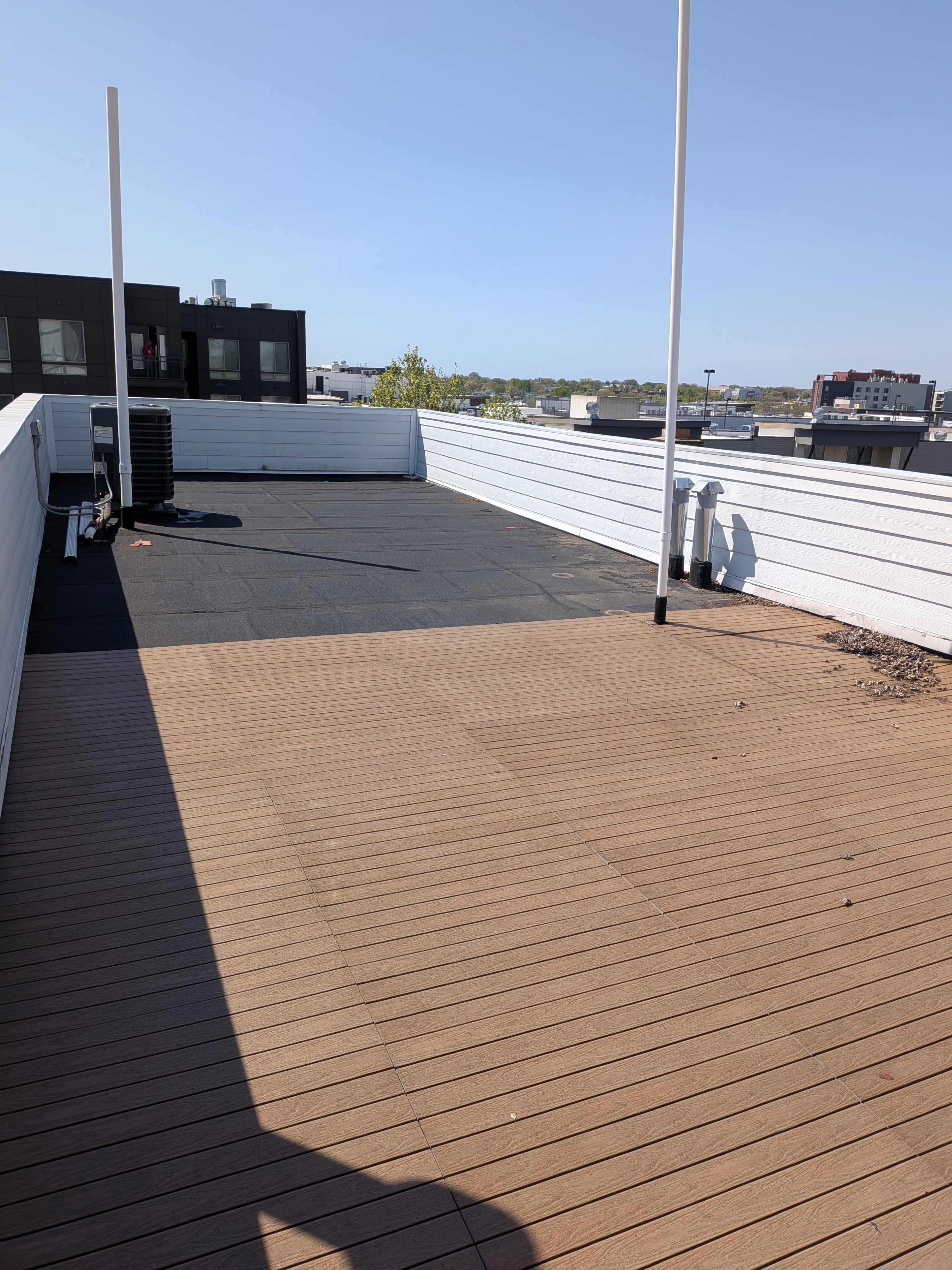 The image shows a rooftop terrace featuring a section of wooden decking and a black surface with a clear blue sky in the background.
