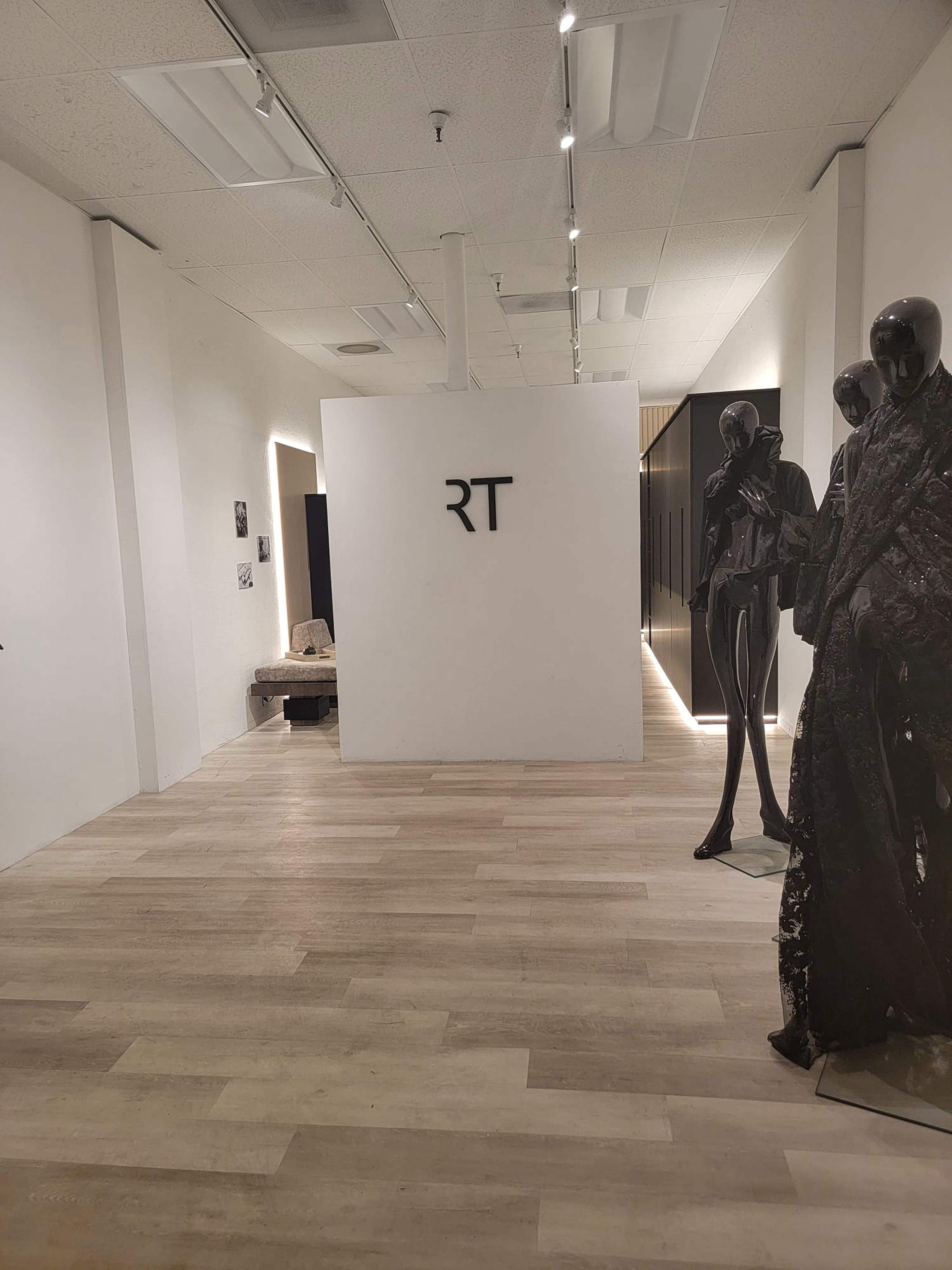 The image shows a modern gallery hallway with a white wall featuring the letters "RT," alongside a sculpture of dark figures and wooden flooring.