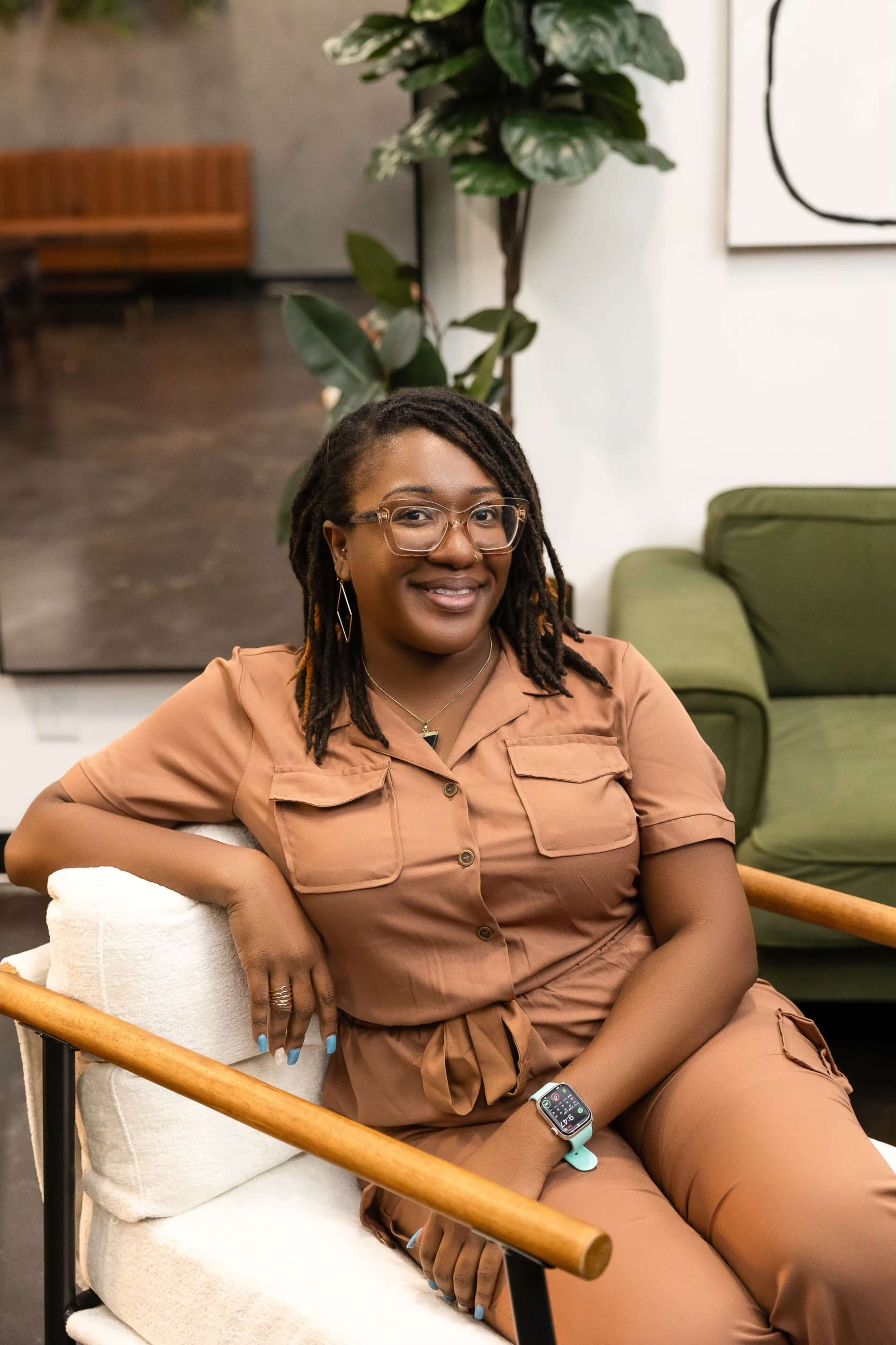A person with glasses and dreadlocks sits comfortably in a modern chair, smiling at the camera in a stylish indoor space.