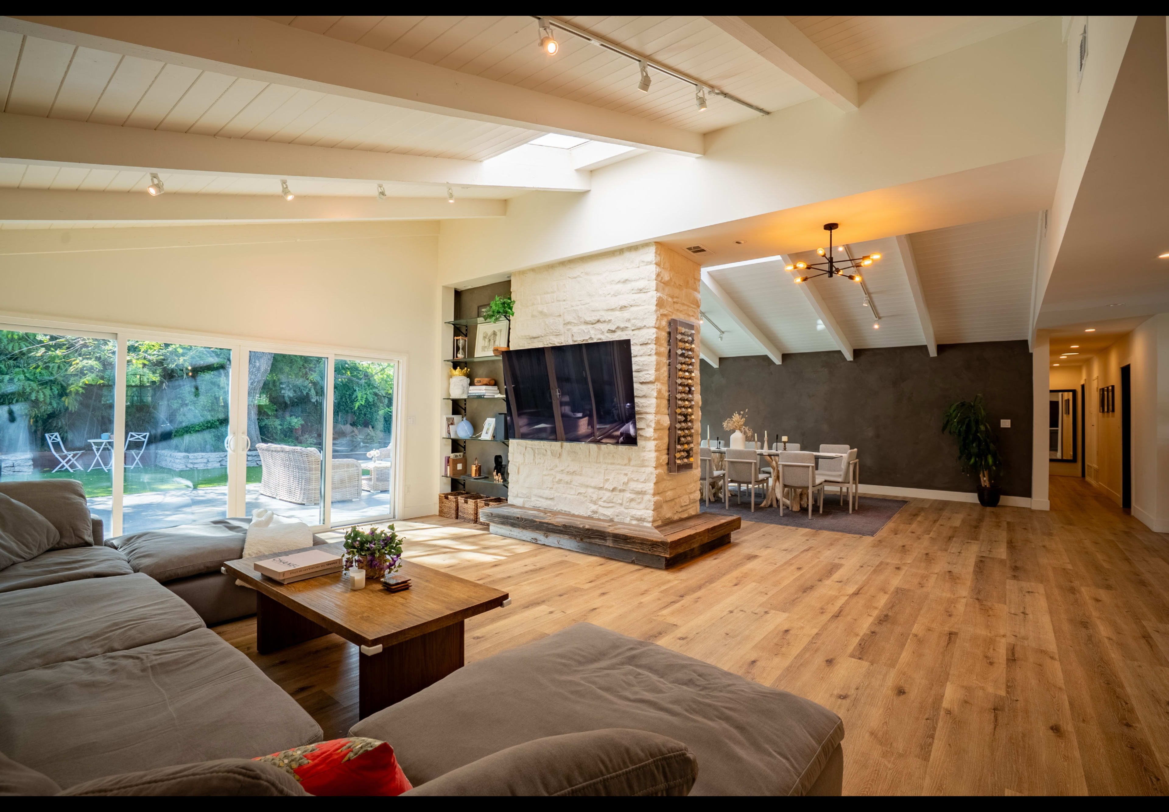 A spacious living room features a large sectional sofa, a wooden coffee table, a TV mounted on a white stone wall, and ample natural light from large windows.