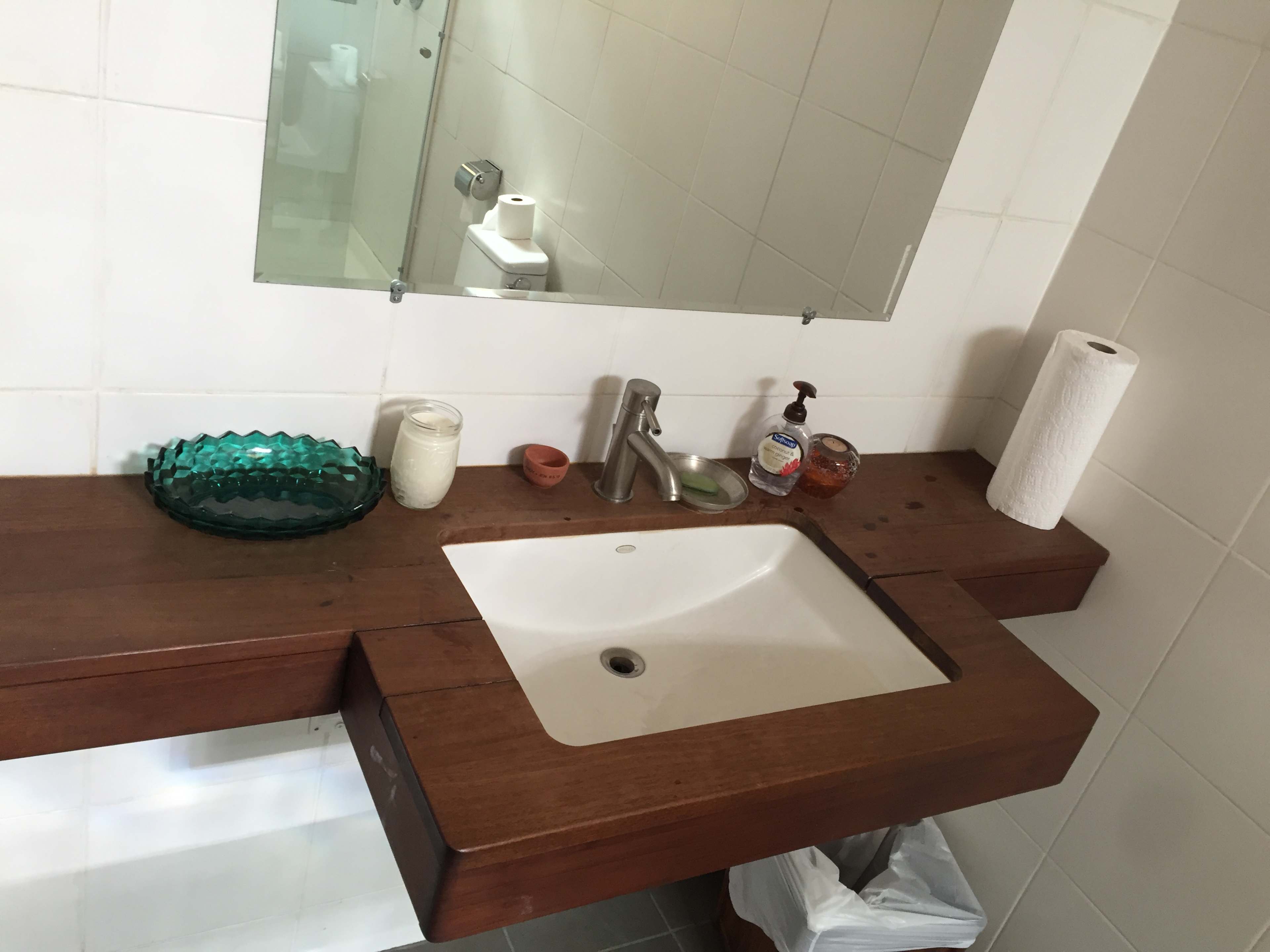 A wooden countertop with a sink, a mirror, and various toiletries beside a roll of paper towels and a trash can.