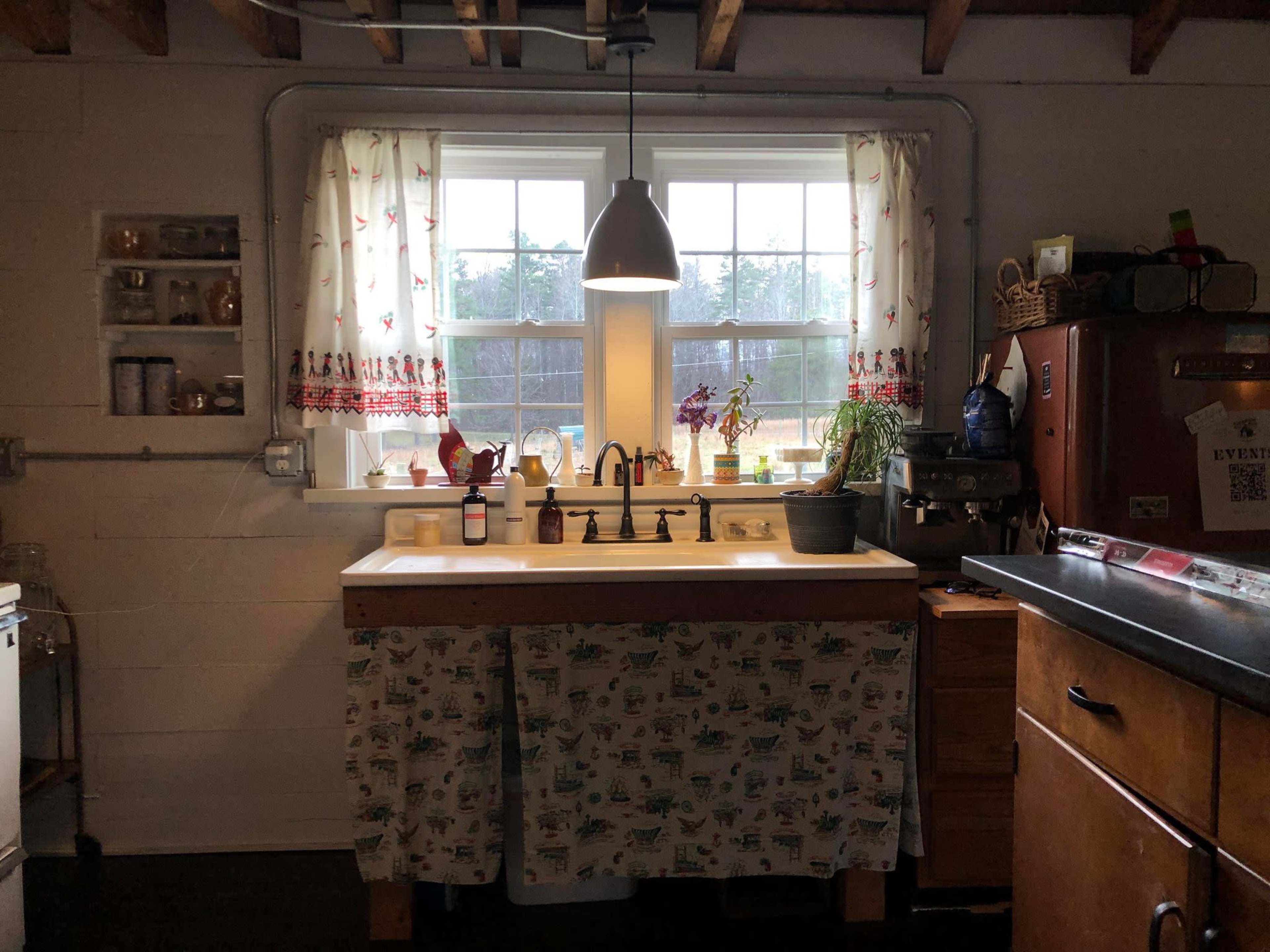 A kitchen with a white sink beneath two windows, a floral curtain, and a mix of modern and vintage appliances.