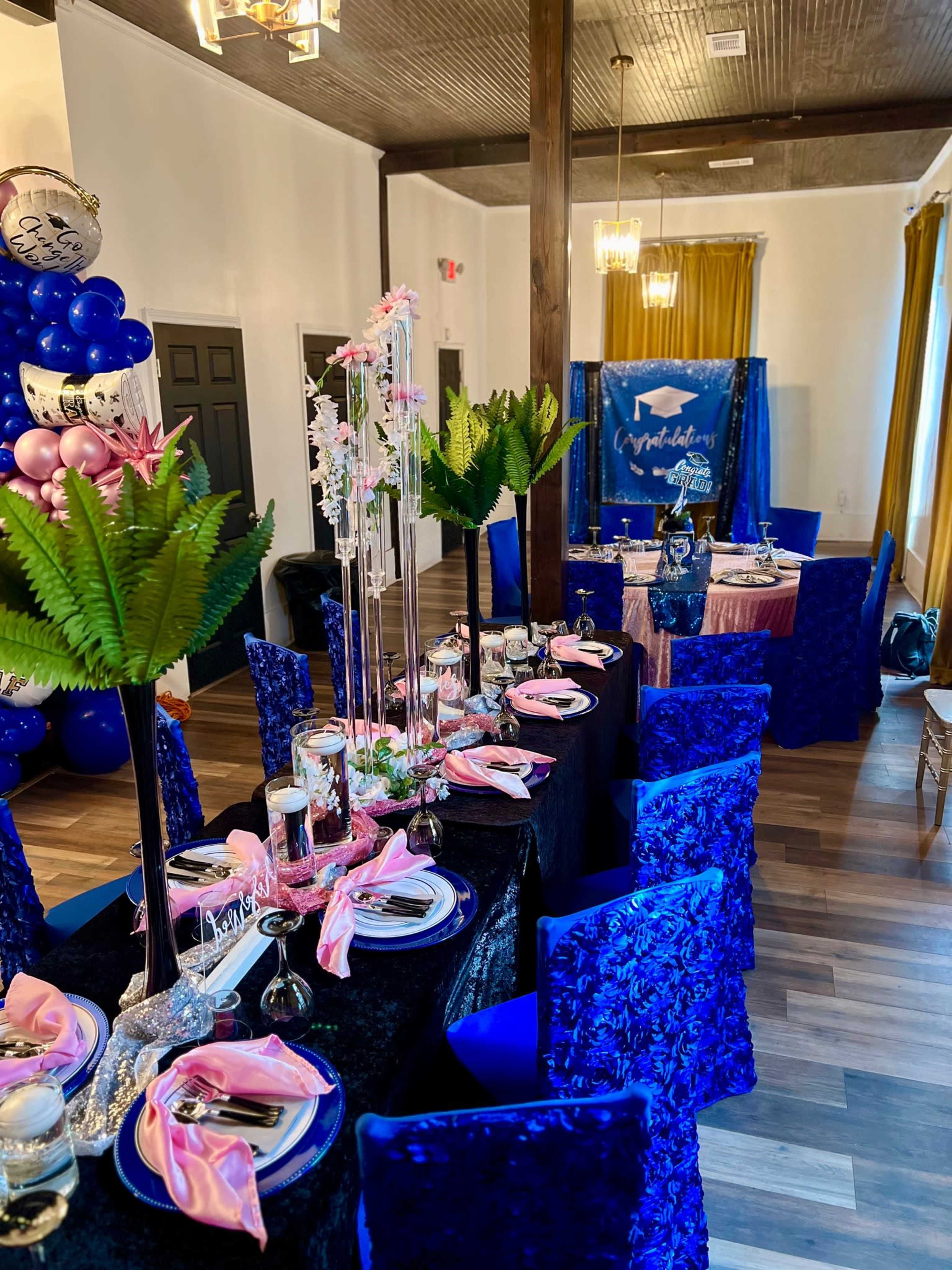 The image shows a decorated banquet hall featuring a long table set with black and blue tablecloths, pink napkins, and floral centerpieces, surrounded by blue chairs, balloons, and a backdrop celebrating a graduation theme.