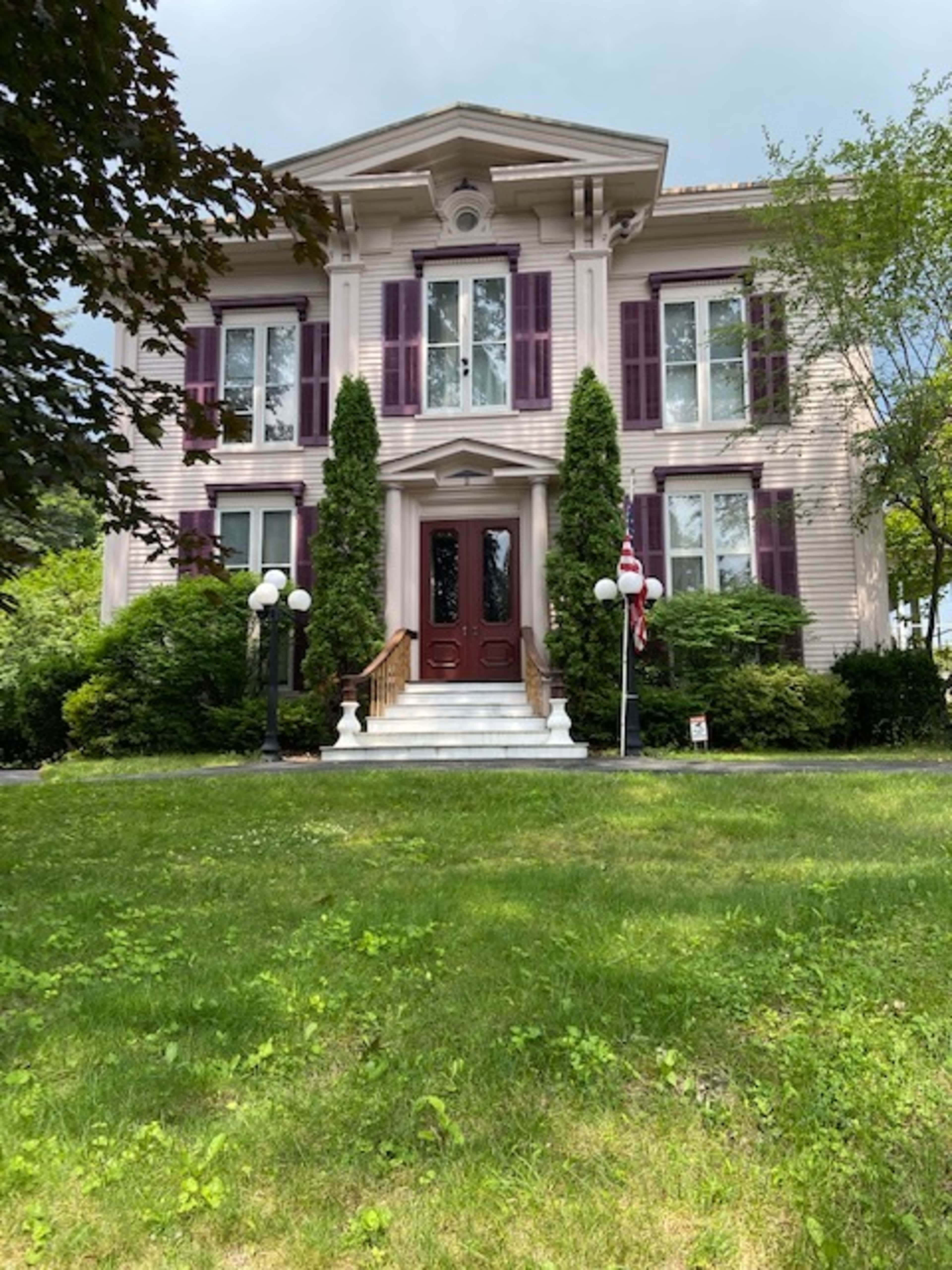A large, two-story house with white siding, purple shutters, and a front porch flanked by tall, green shrubs.
