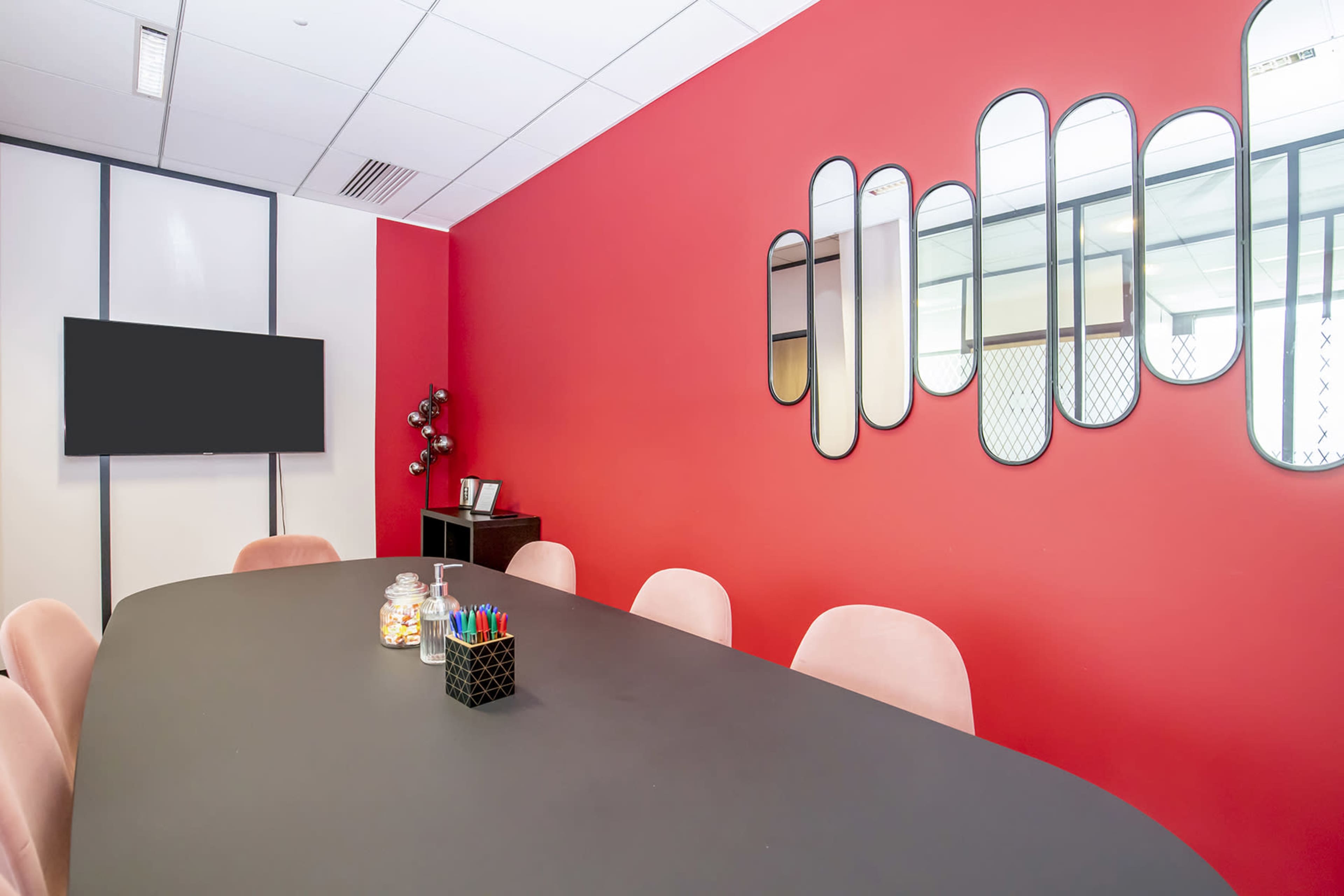 A modern conference room features a black oval table, pink chairs, a television on the wall, and decorative mirrors on a red accent wall.