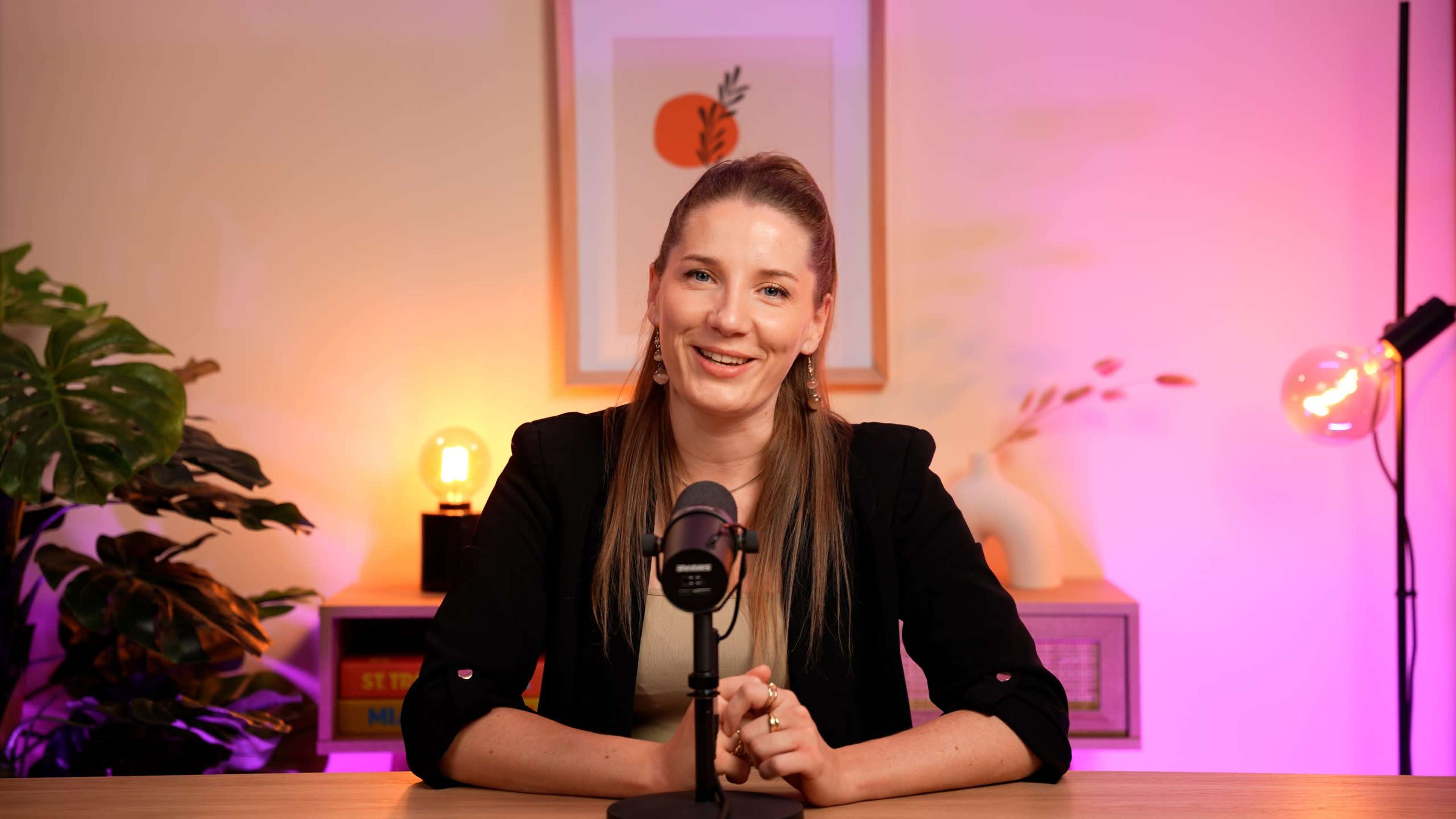 A woman with long hair is sitting at a table in front of a microphone, smiling, with colorful lighting and decorative elements in the background.