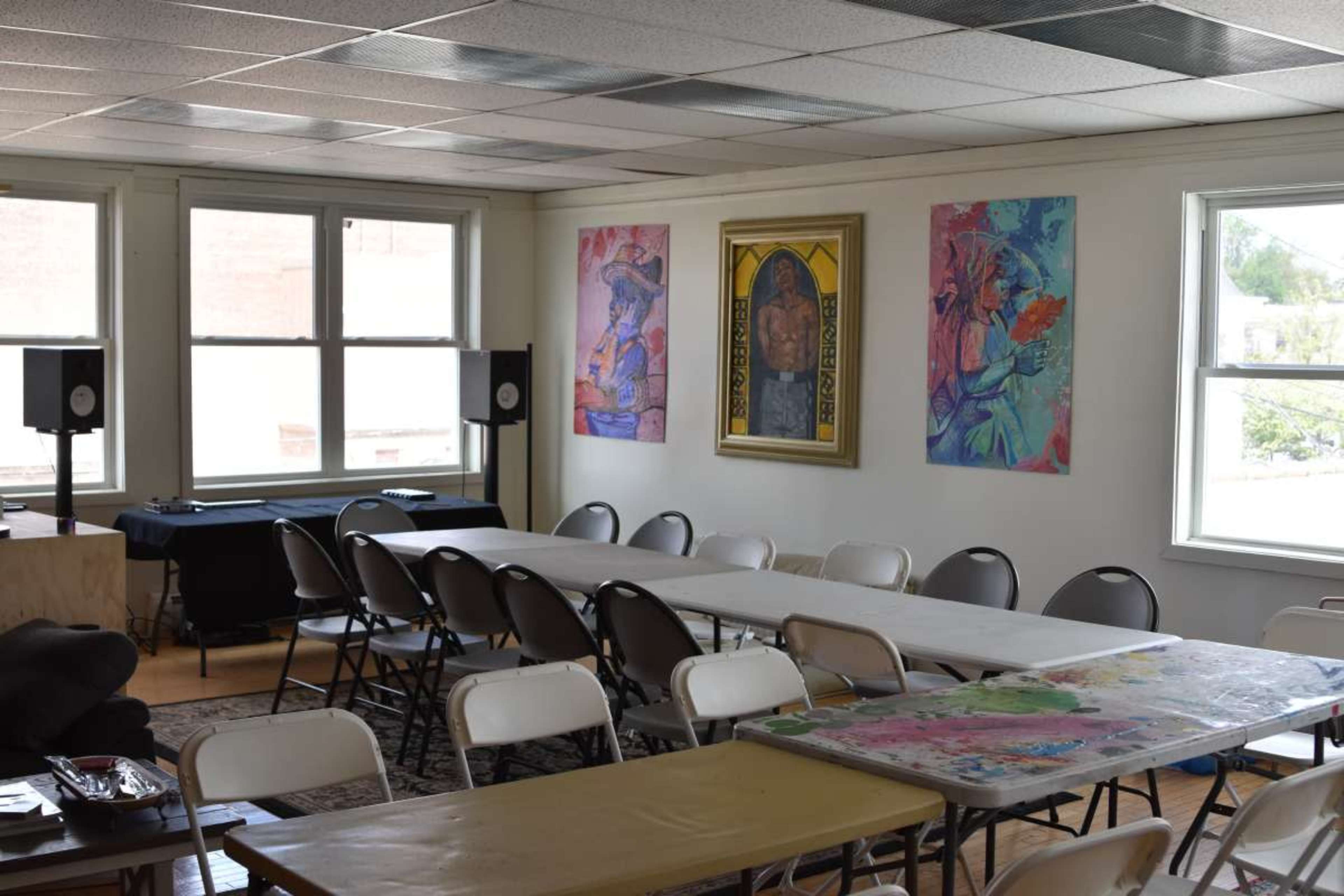 The image shows a spacious room with rows of white chairs and tables, decorated with colorful artwork on the walls and audio equipment in the corners.