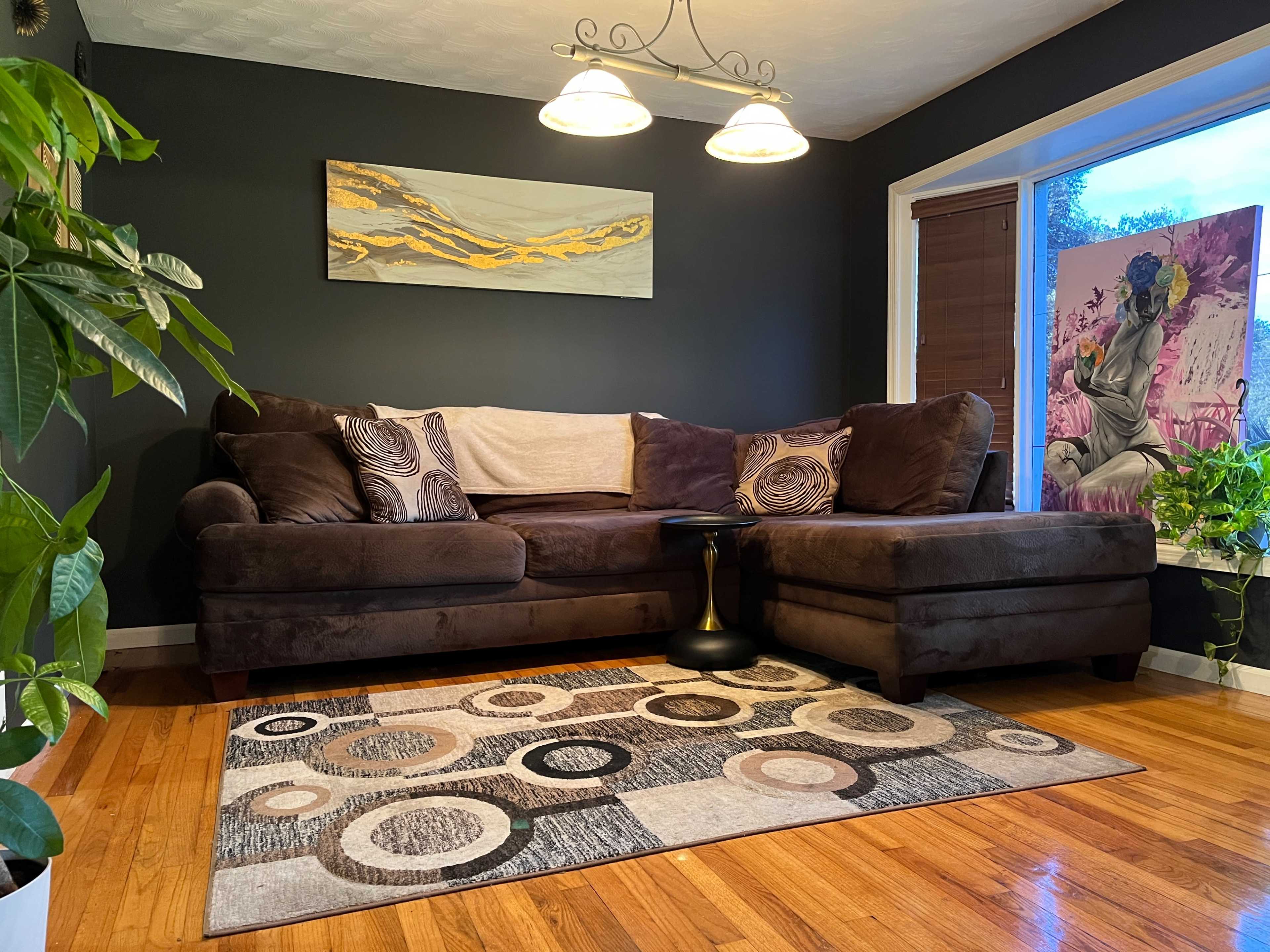The image shows a living room with a dark wall, a brown sectional sofa, a round coffee table, a patterned area rug, and a large window displaying a plant and artwork.