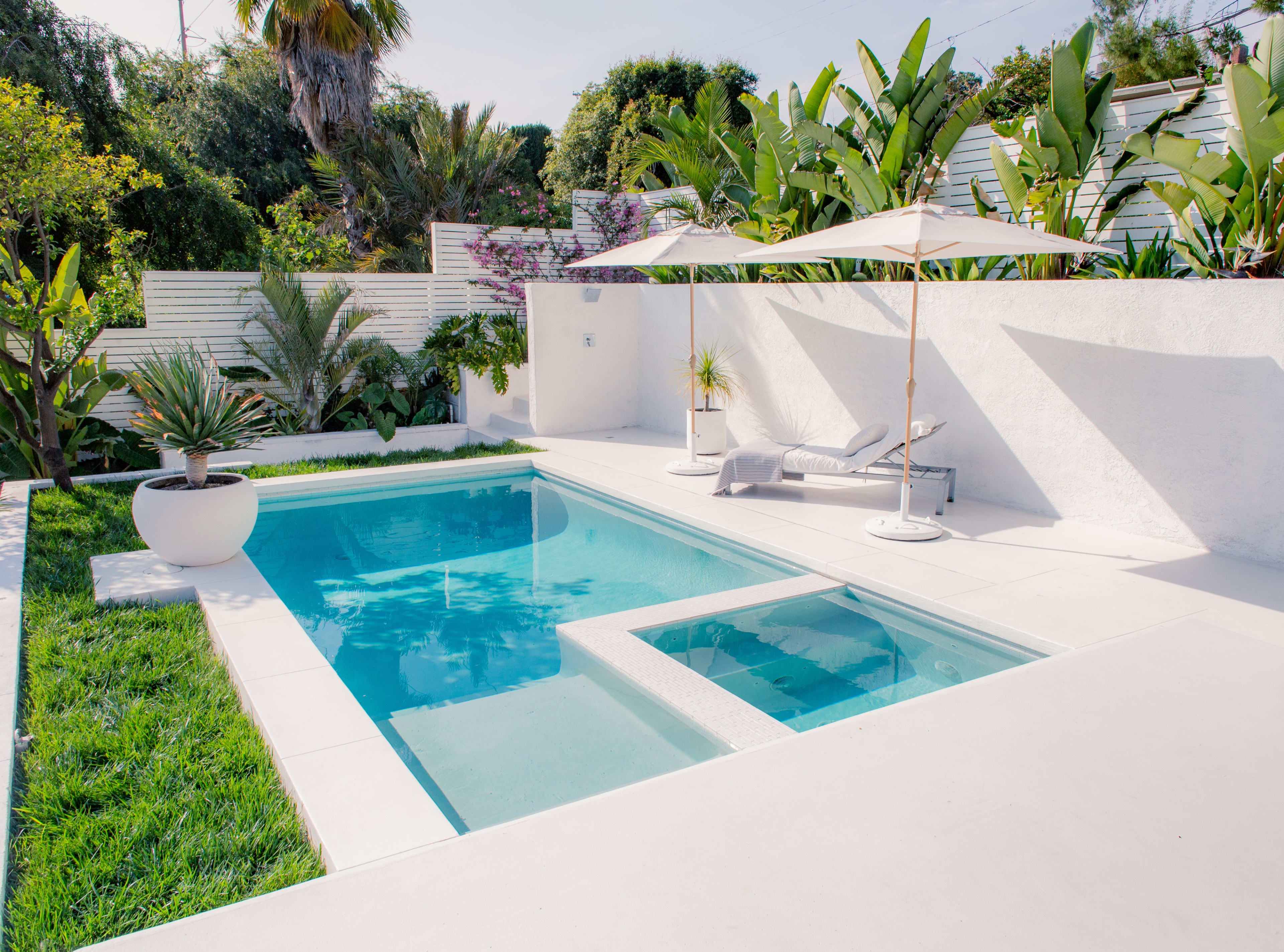 A rectangular swimming pool surrounded by white concrete surfaces, tropical plants, and a pair of lounge chairs with umbrellas.
