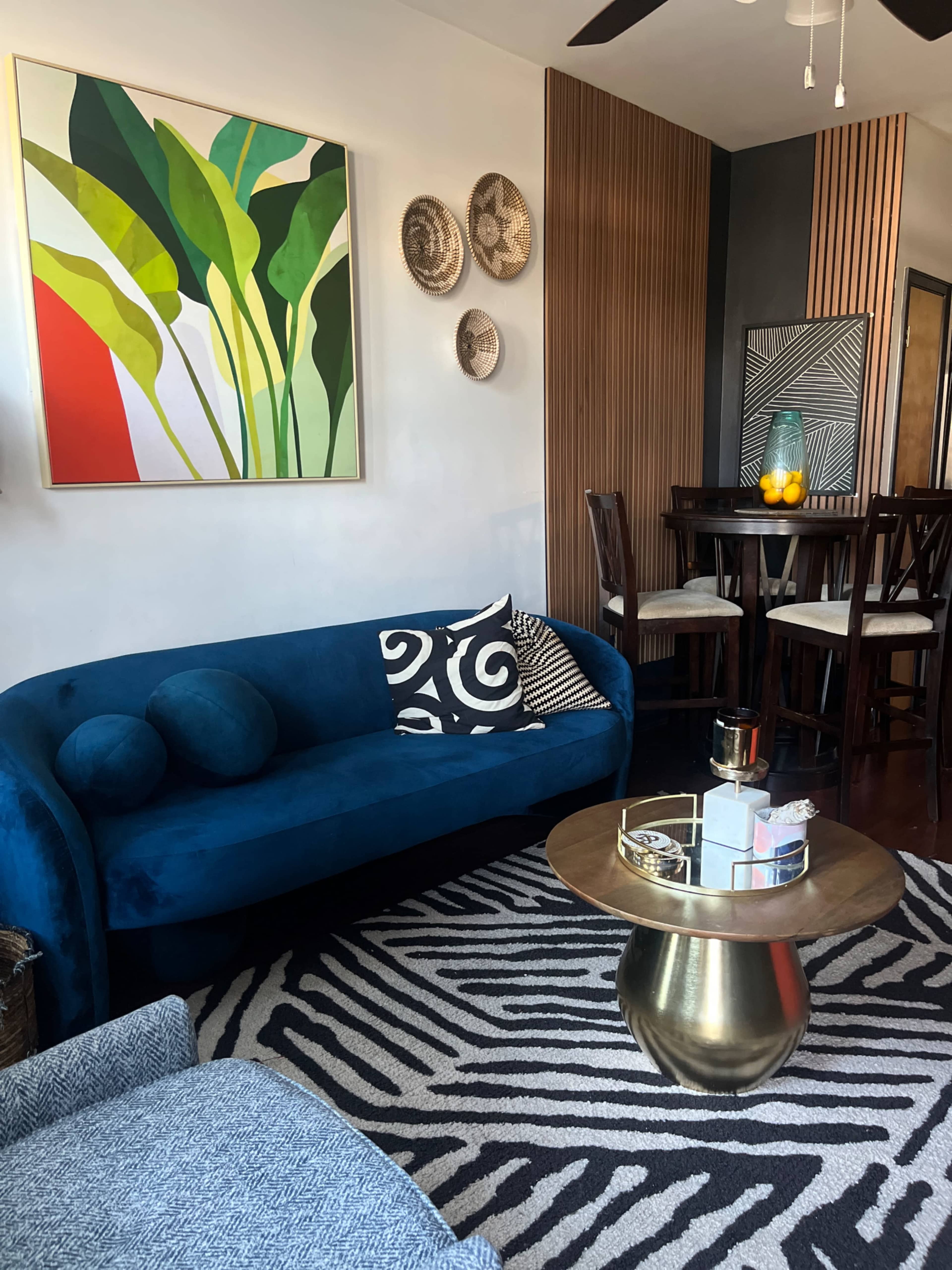 The image shows a modern living room featuring a blue sofa, a round gold coffee table, a zebra-striped rug, a dining area with wooden chairs, and vibrant wall art.