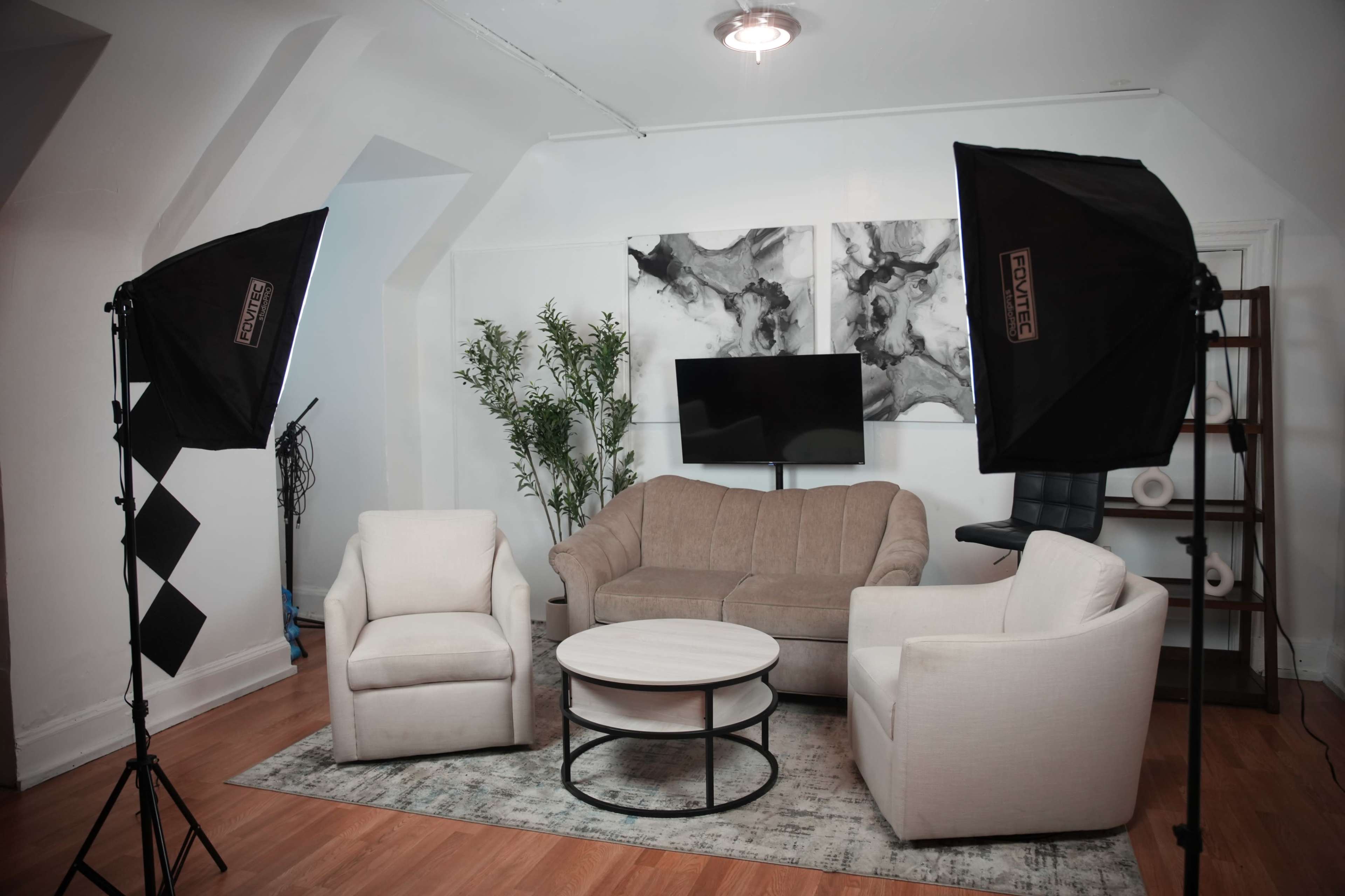 A living room setup features a beige sofa and two white armchairs surrounding a round coffee table, with a television on a stand and lighting equipment in the corners.