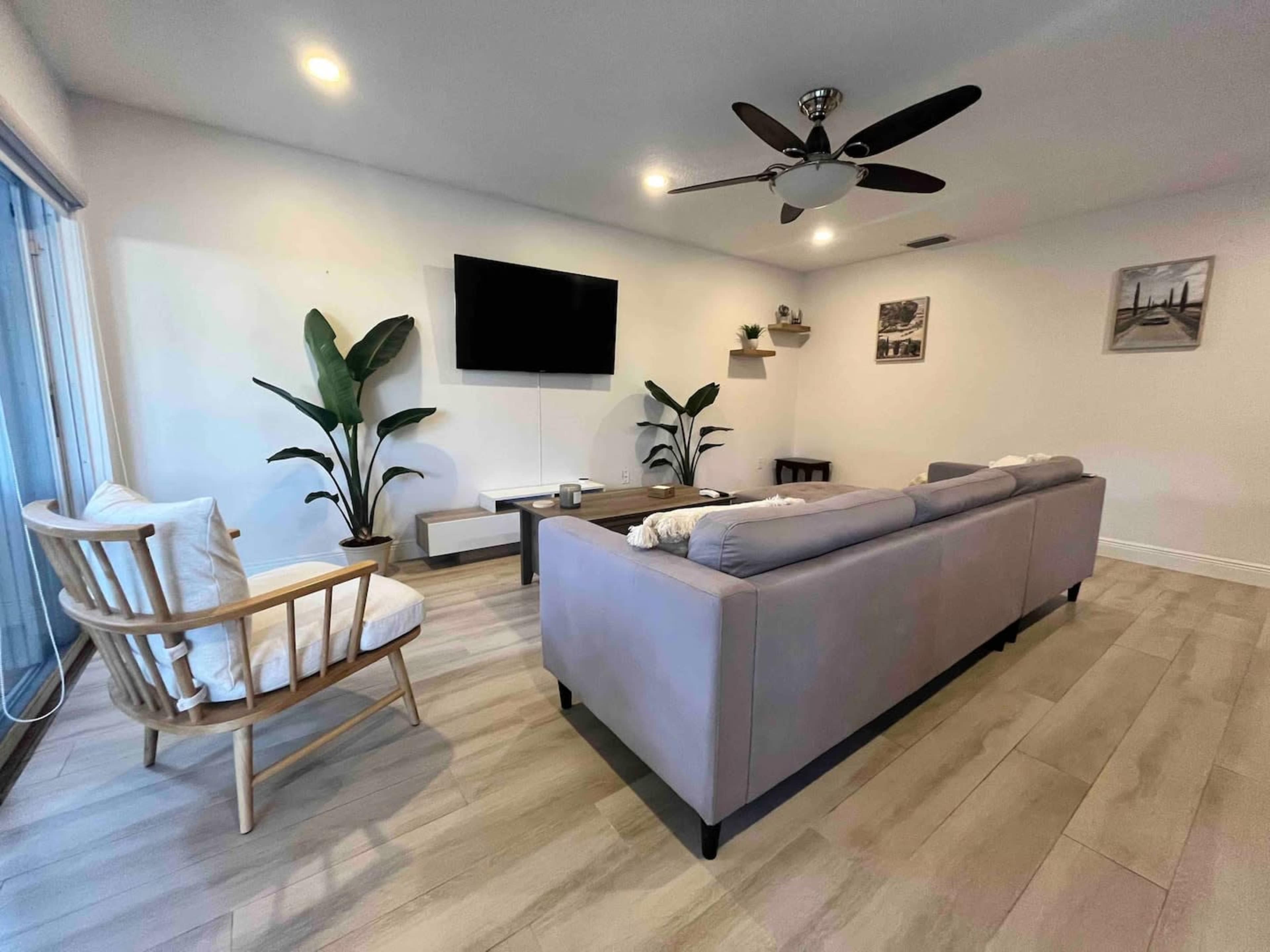 The image shows a modern living room with a gray sofa, a wooden chair, two potted plants, a wall-mounted TV, and a light-colored floor.