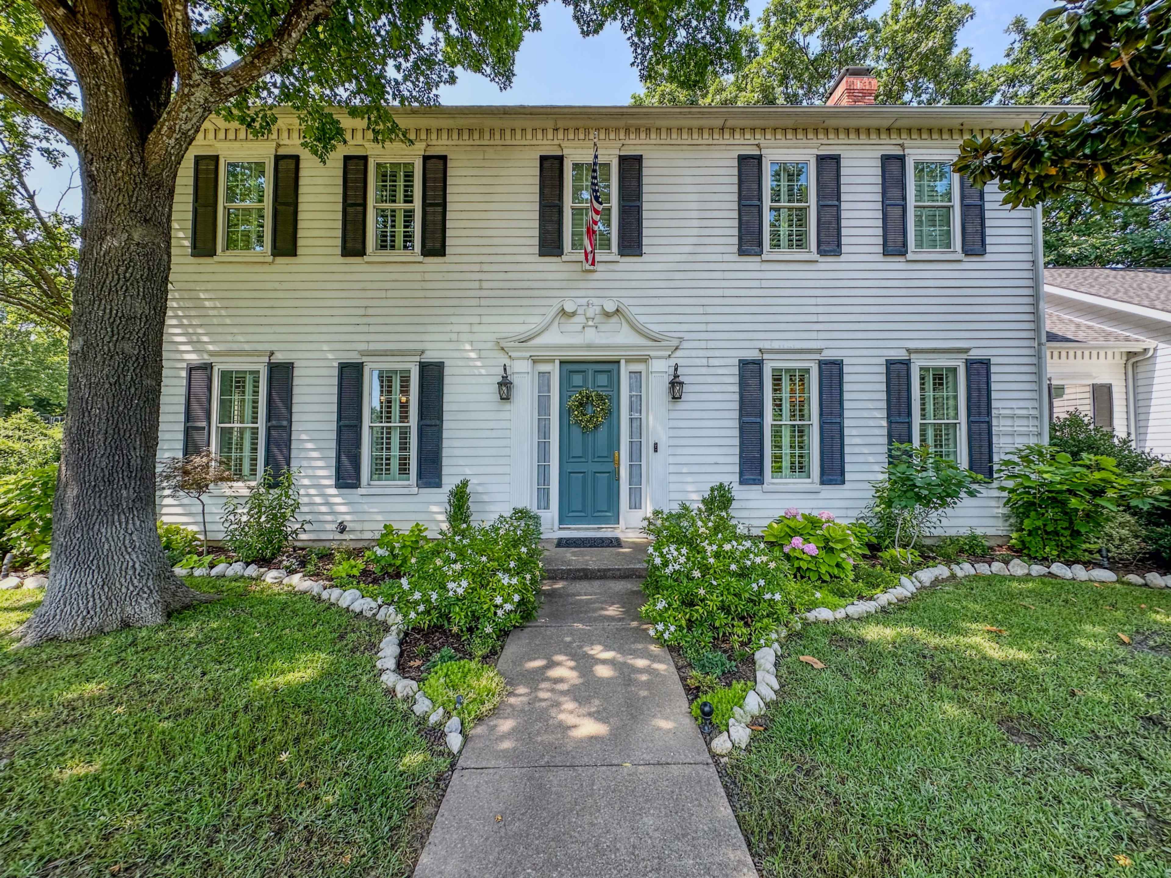 The image shows a two-story white colonial-style house with black shutters, a blue front door, and a manicured garden pathway leading up to it.