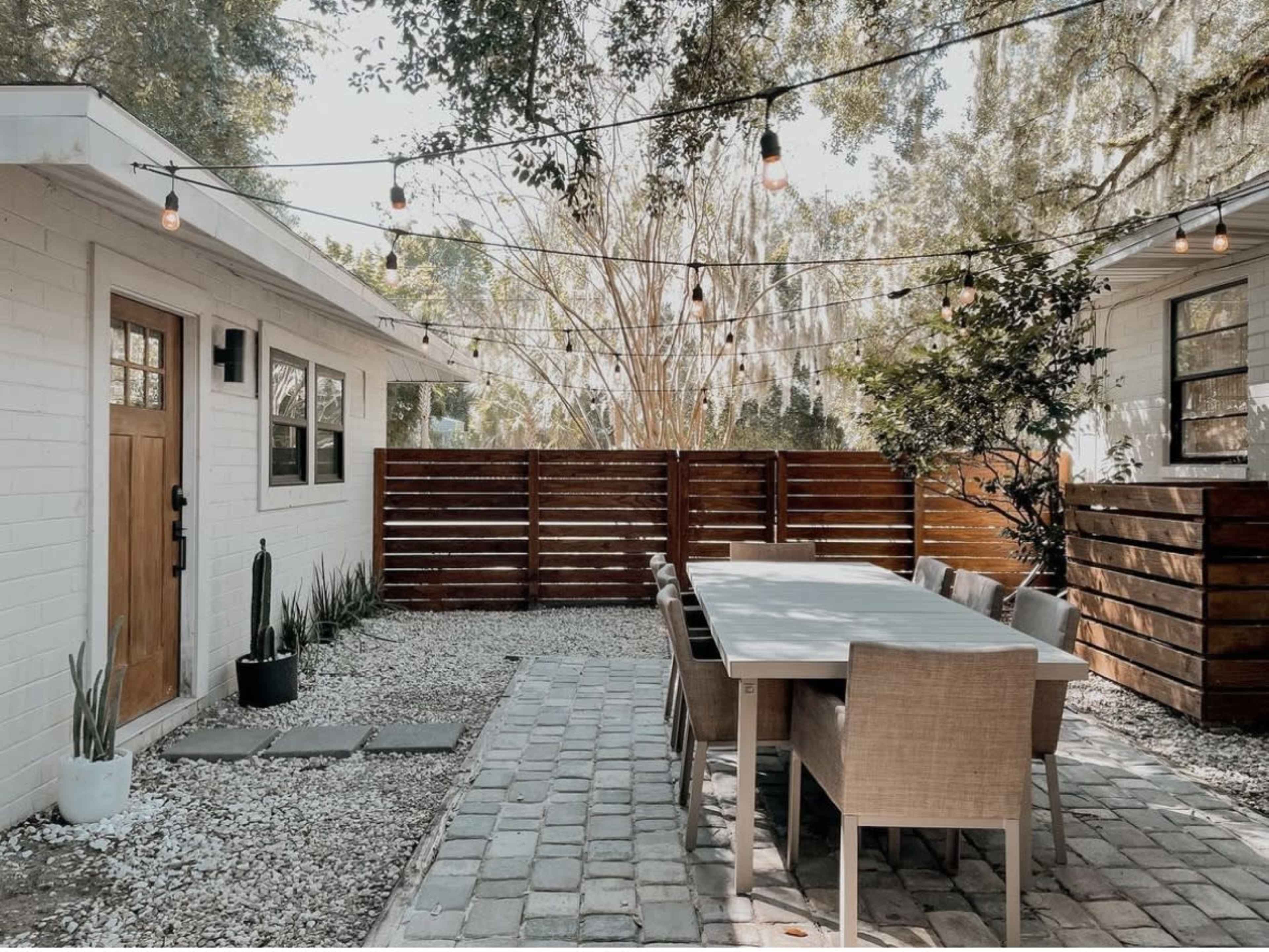 A spacious outdoor dining area features a long table surrounded by chairs, set among stone paths and surrounded by wooden fencing with hanging lights overhead.