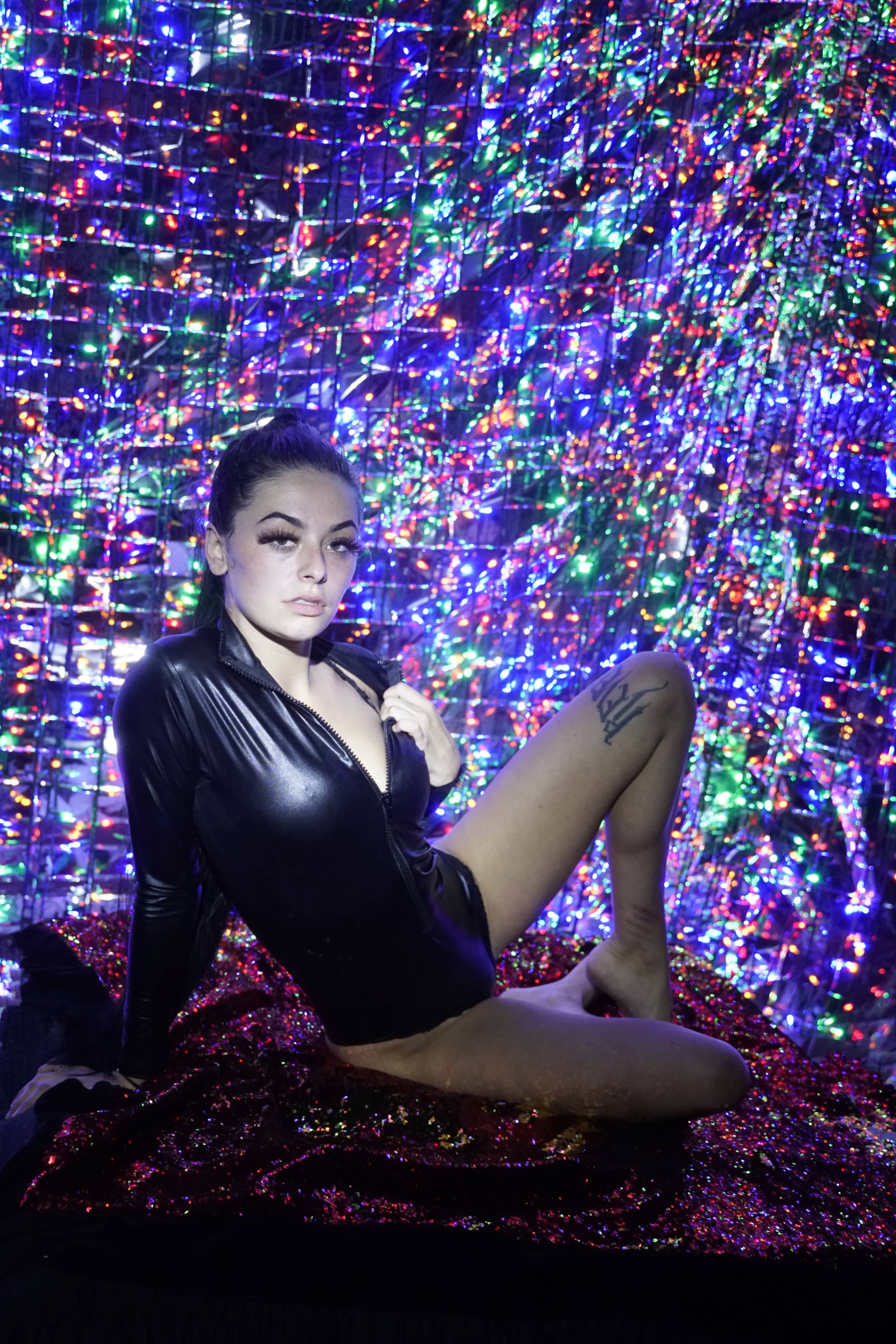 A person poses in a black outfit against a backdrop of colorful, shimmering lights.