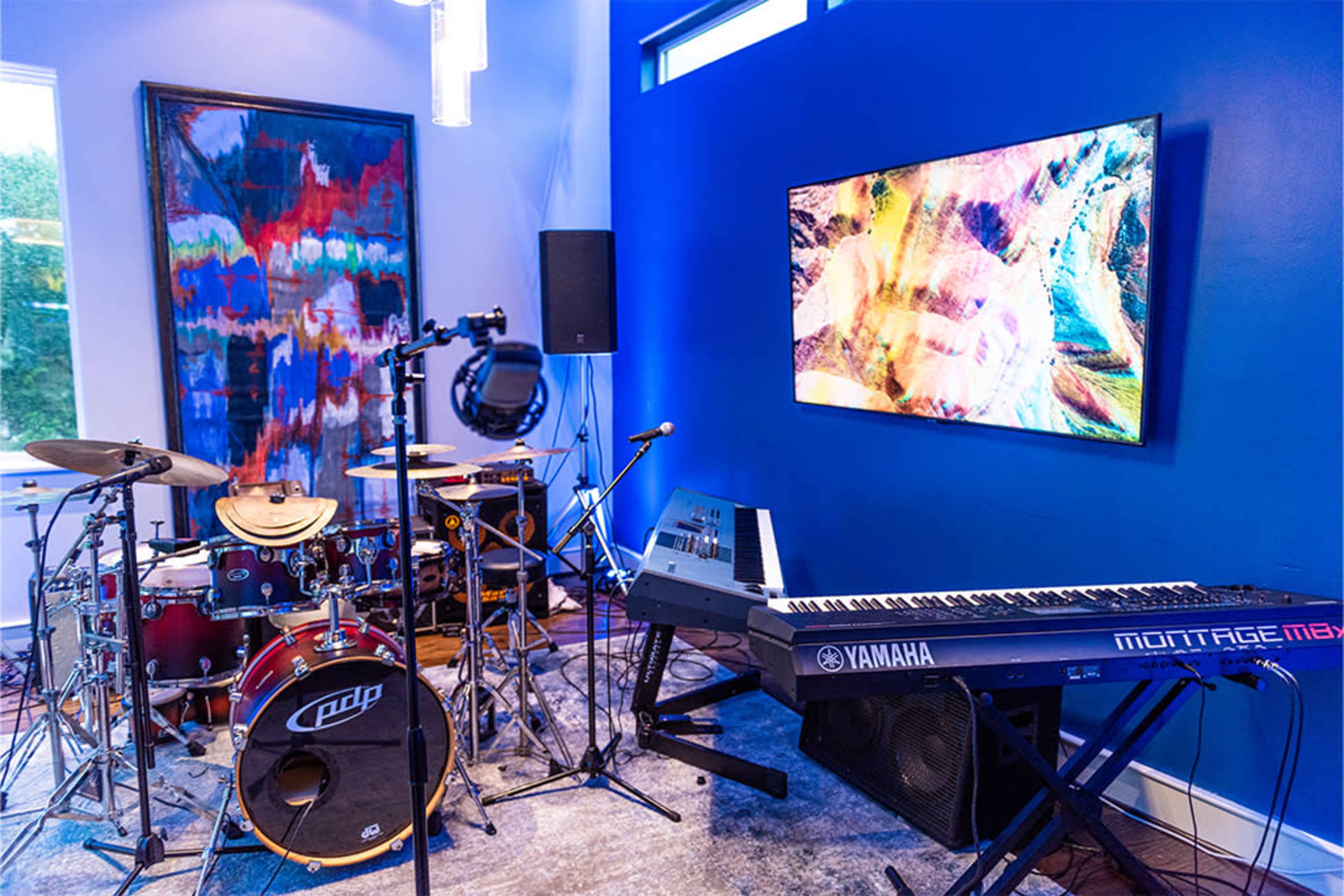 The image shows a music room equipped with a drum set, a keyboard, and a large wall-mounted television displaying colorful graphics.