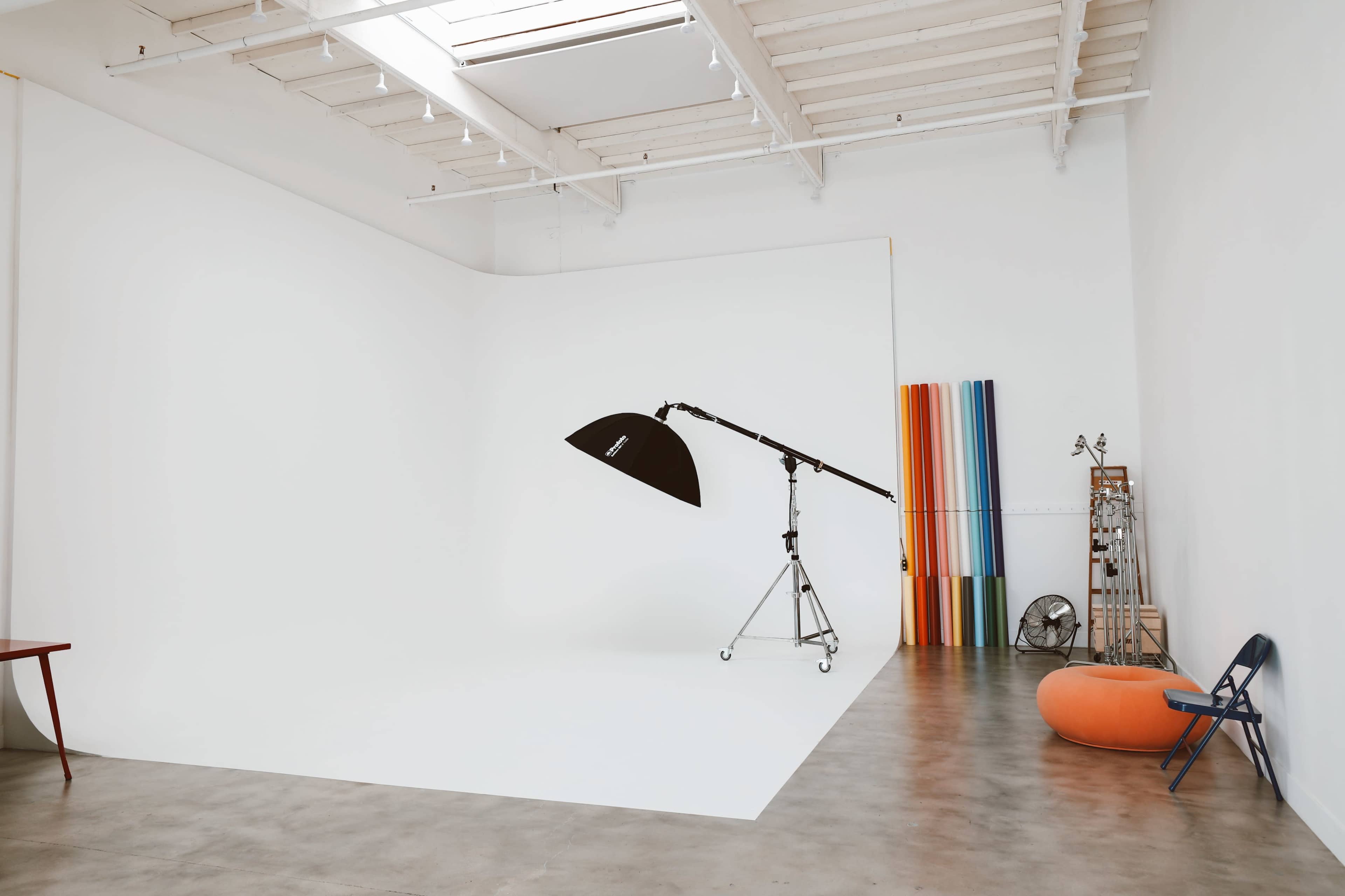 A spacious photography studio features a large white backdrop, a mounted light stand, and colorful backdrops neatly arranged along the wall.