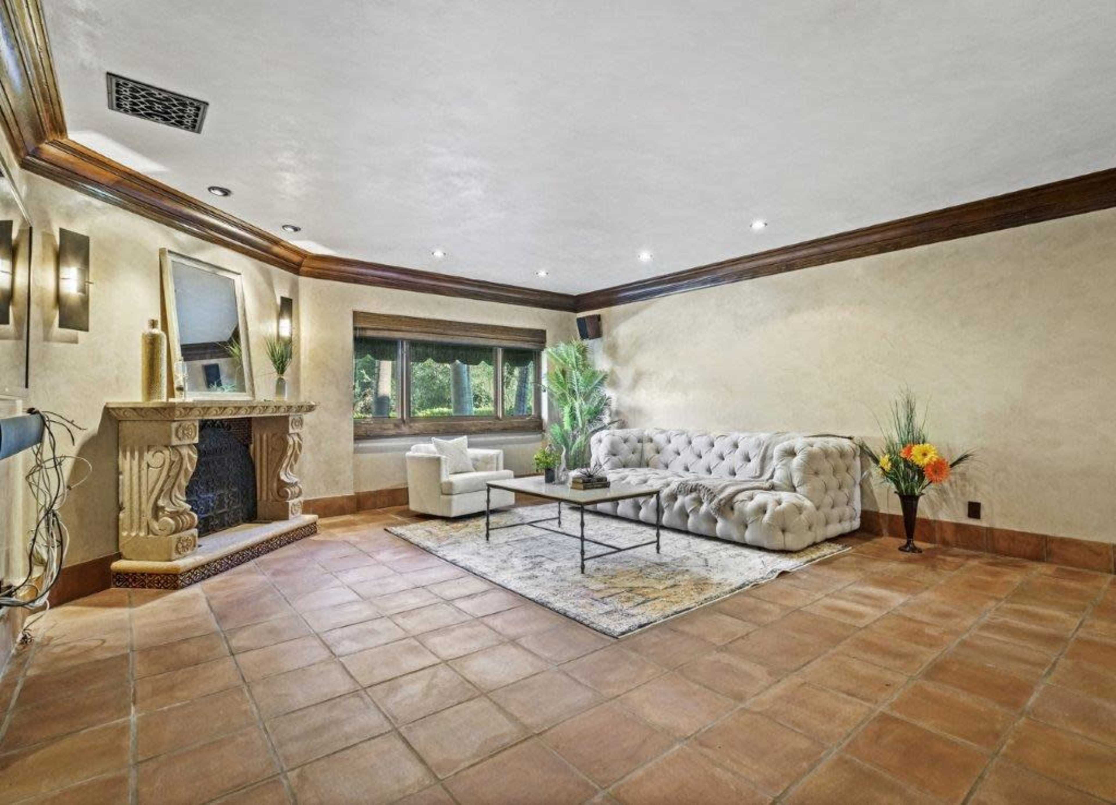 The room features a light-colored wall with a decorative fireplace, a large tufted sofa, a coffee table, and a large window allowing natural light to enter.