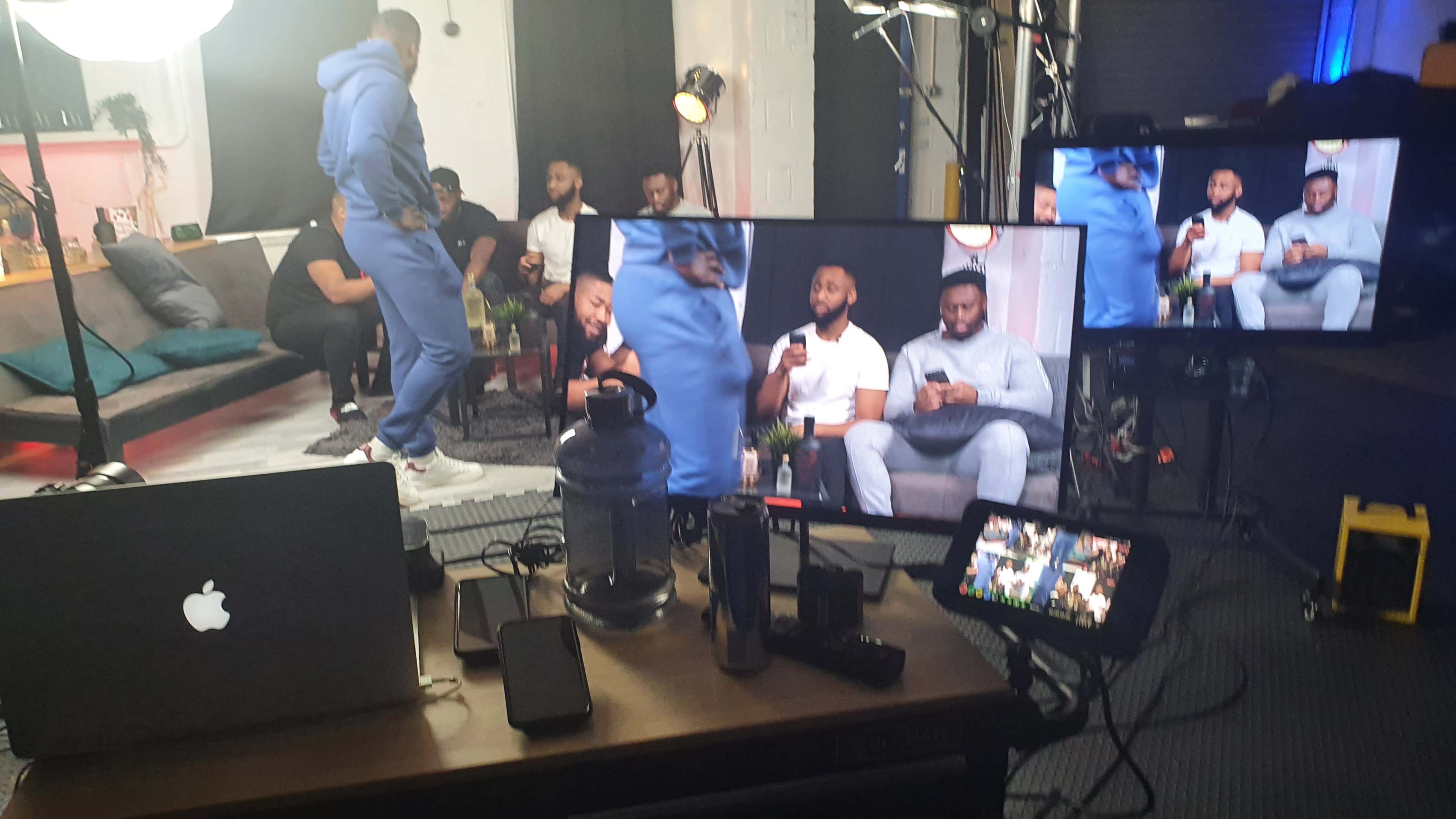 A filming setup with multiple monitors displaying a group of men sitting on a couch, while various equipment and devices are arranged on a table in the foreground.