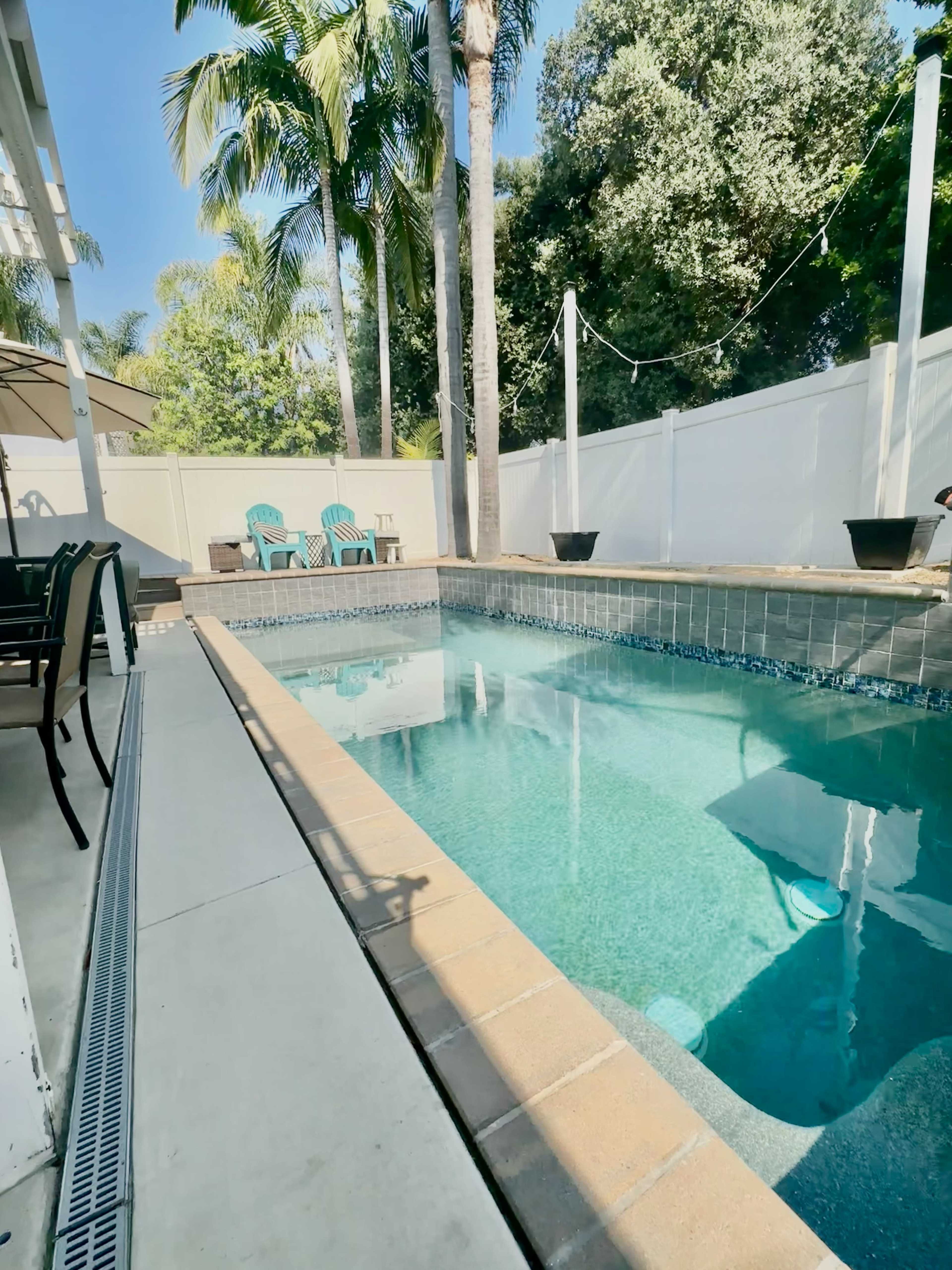 A clear swimming pool is surrounded by palm trees and white fencing, with two blue chairs visible on the patio.