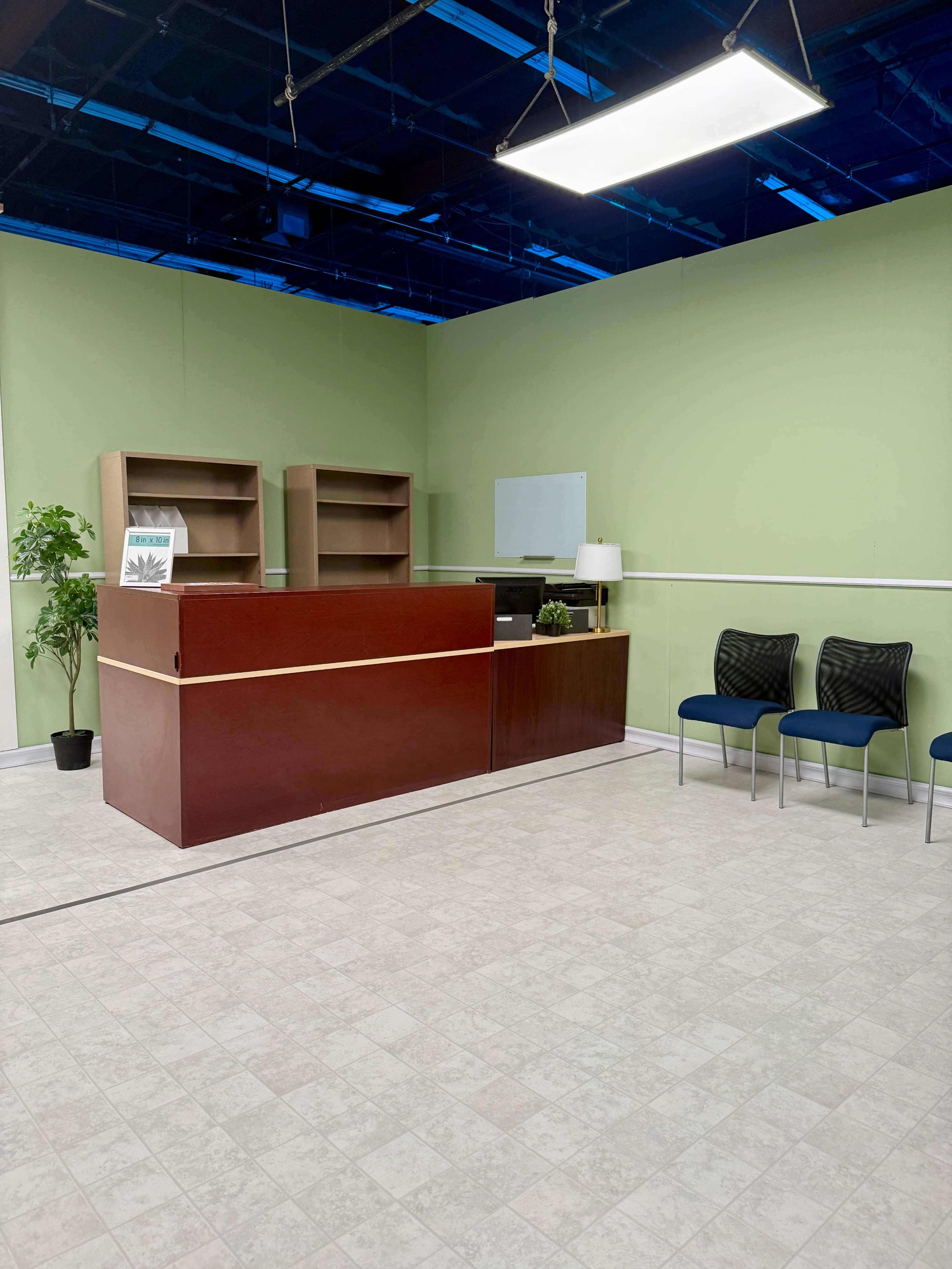The image shows a minimalist reception area with a wooden reception desk, a small lamp, a few chairs, and green walls.