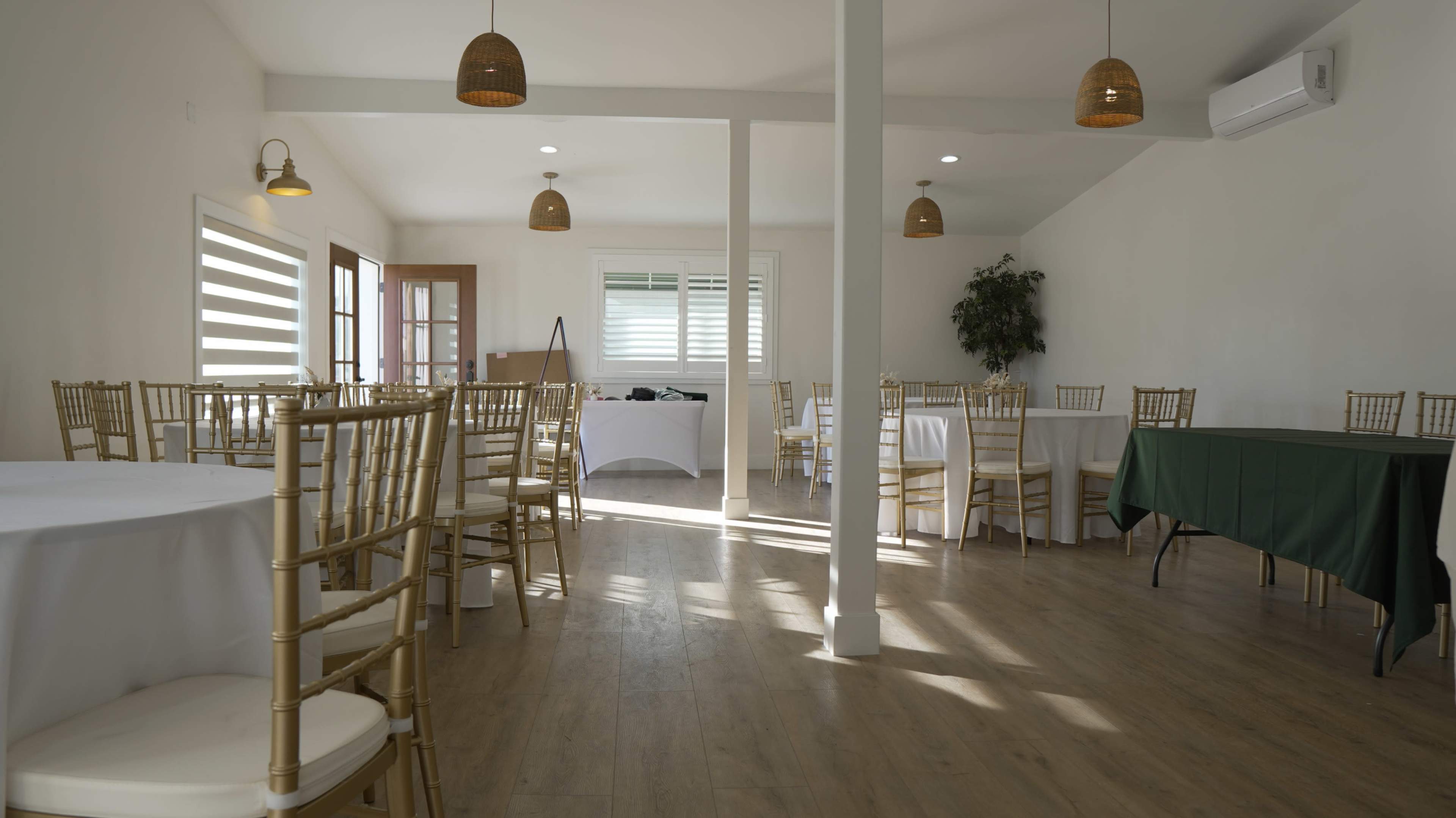 An indoor event space features gold chiavari chairs arranged around tables with white linens, illuminated by natural light streaming through windows.