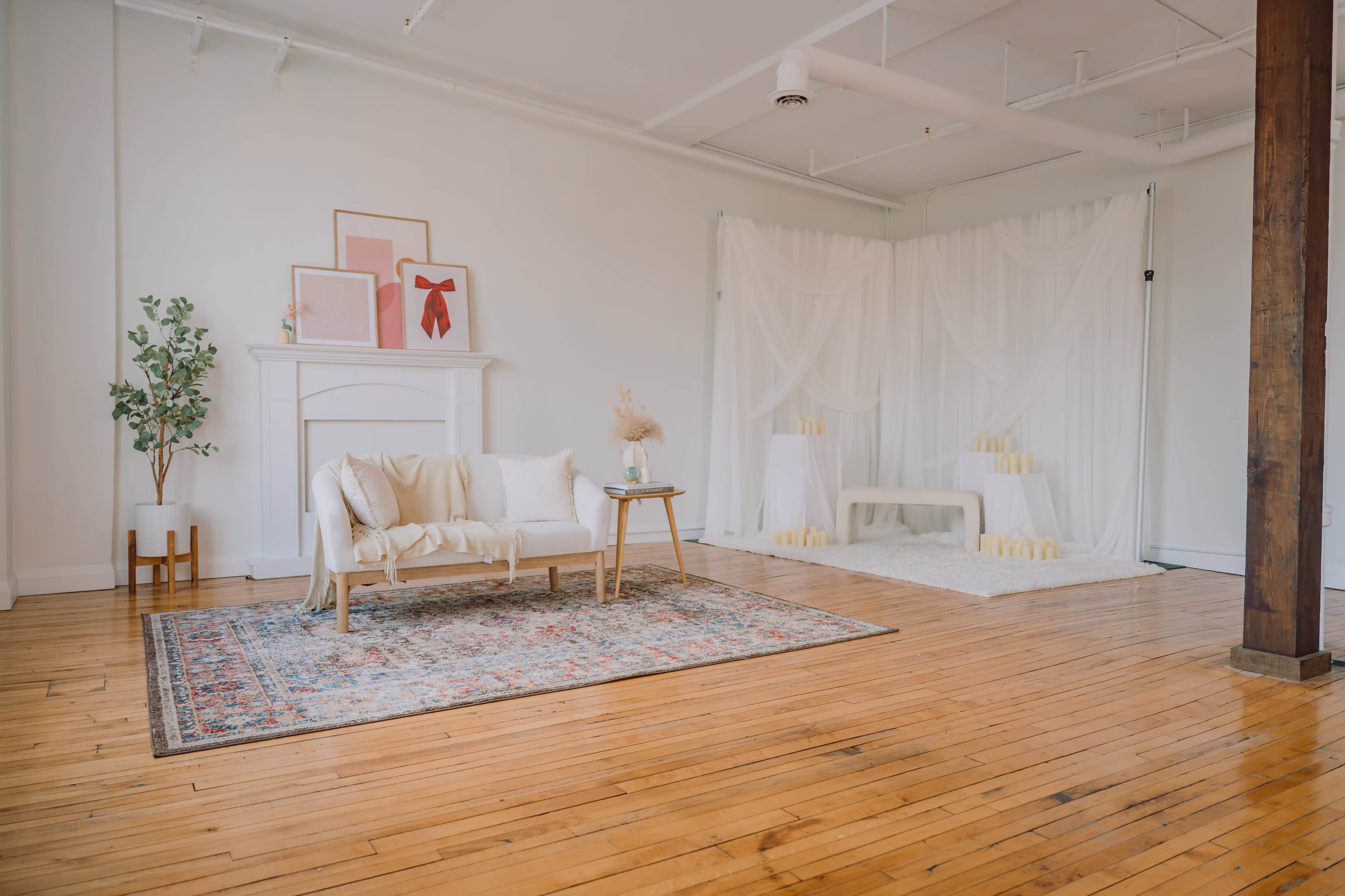The image shows a spacious room with a light-colored wooden floor, featuring a minimalist seating area with a couch, a small table, and decorative elements including plants and soft backdrops.
