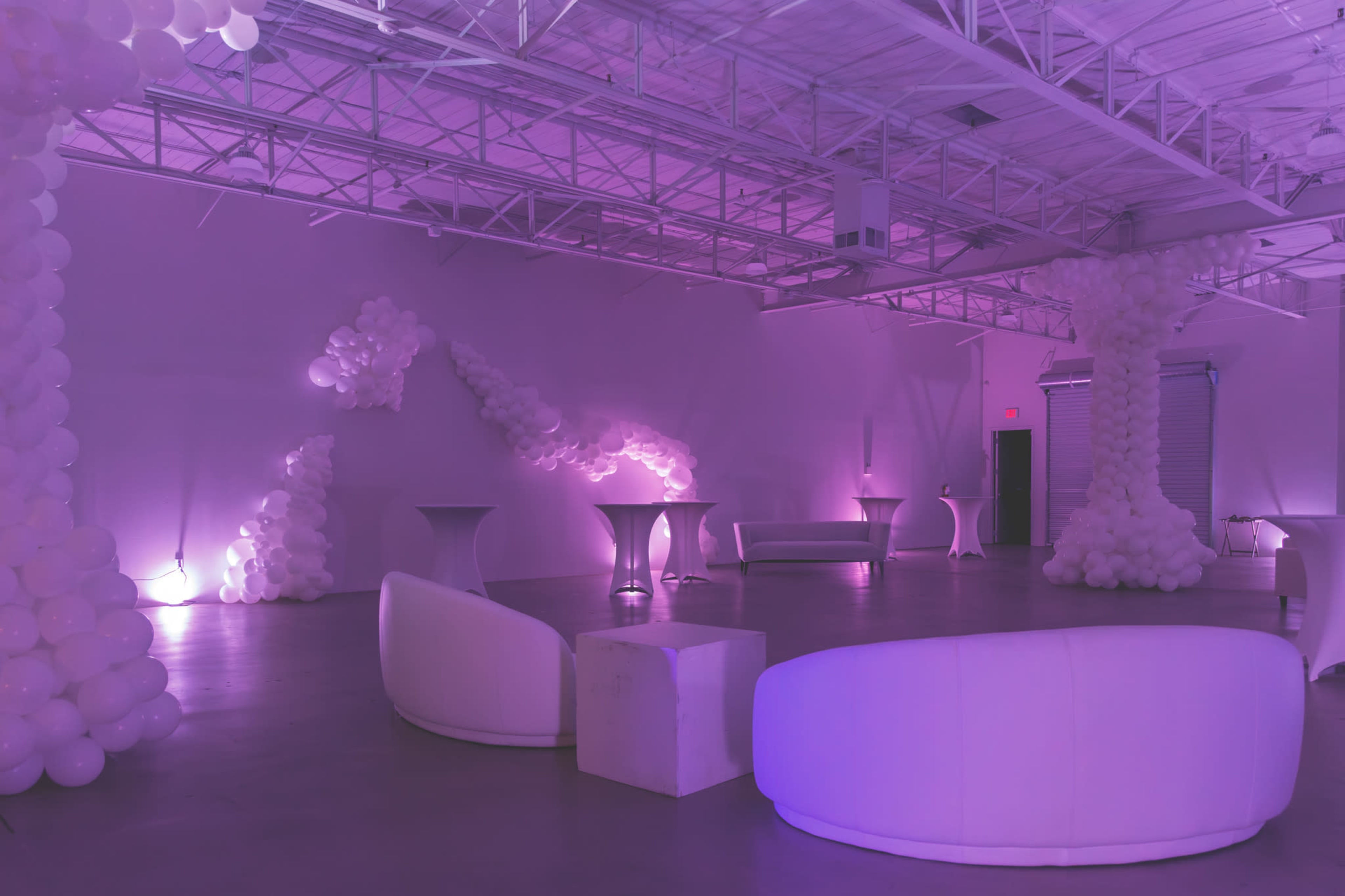 A spacious, softly lit event room features white balloon decorations and modern furniture arranged around the area.