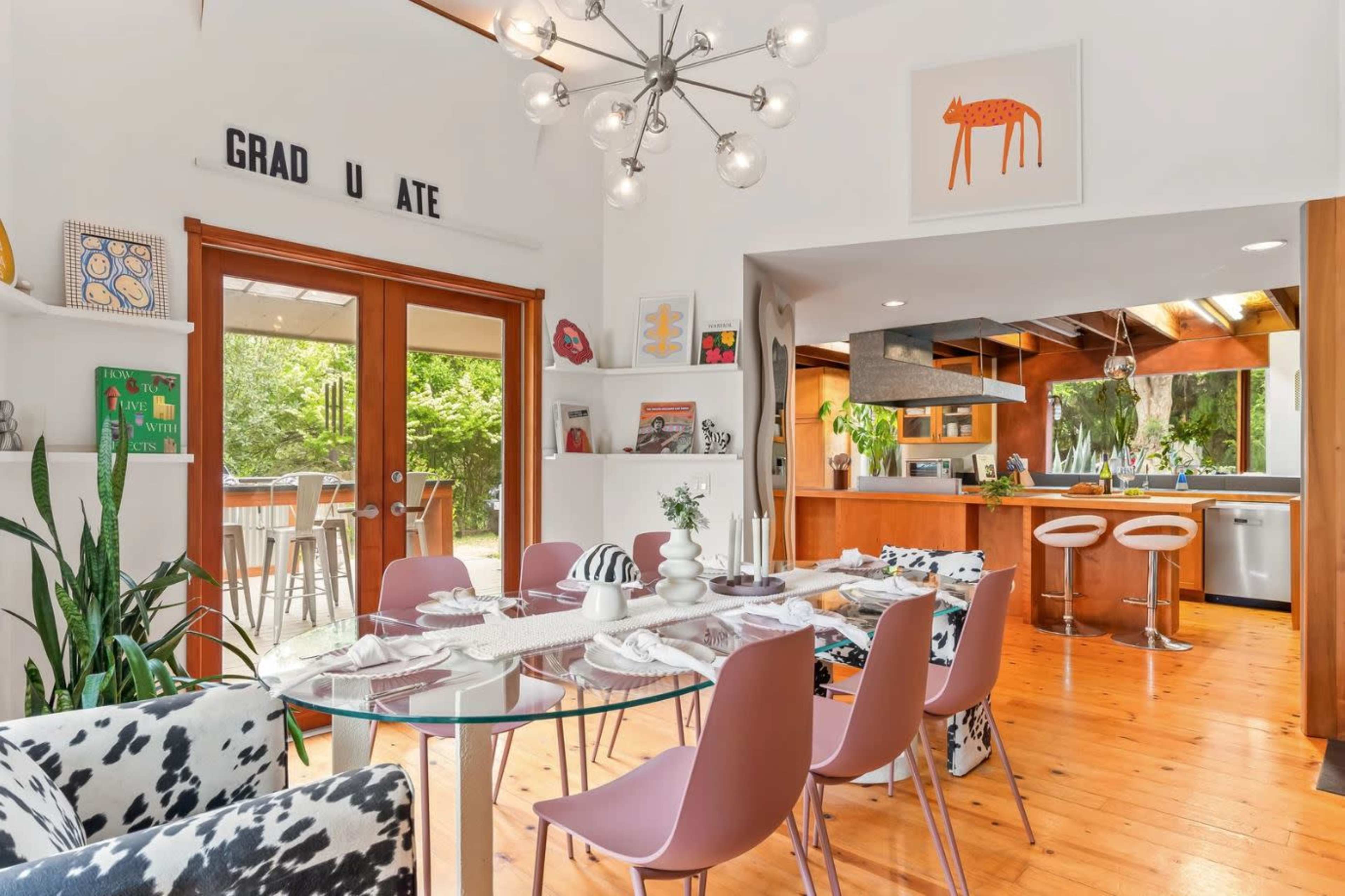 A modern dining area features a glass table surrounded by pink chairs, with an open kitchen visible in the background and large windows letting in natural light.