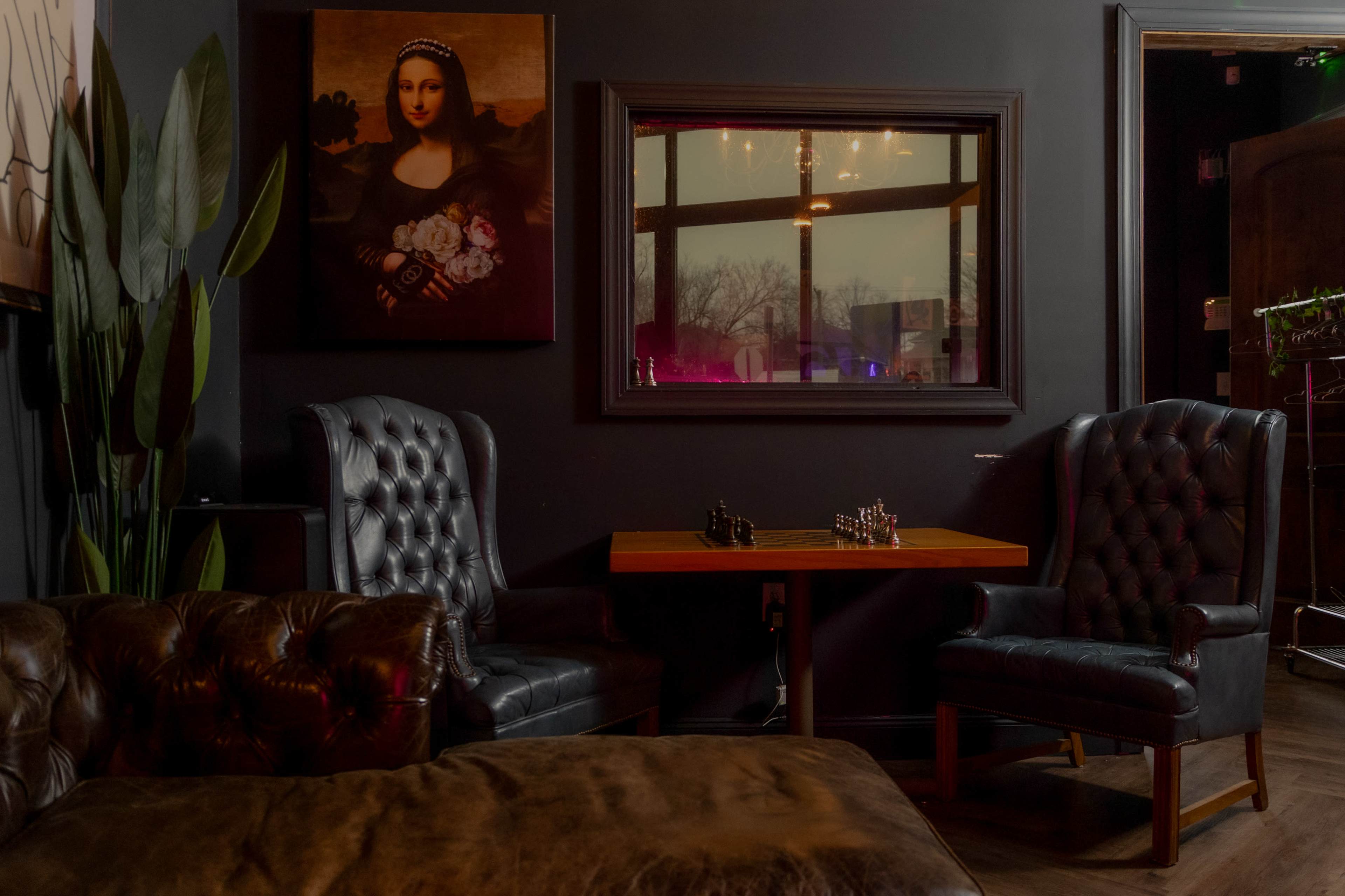 A cozy lounge area with dark walls, featuring a large portrait of a woman, a small wooden table with a chess set, and two tufted leather armchairs.