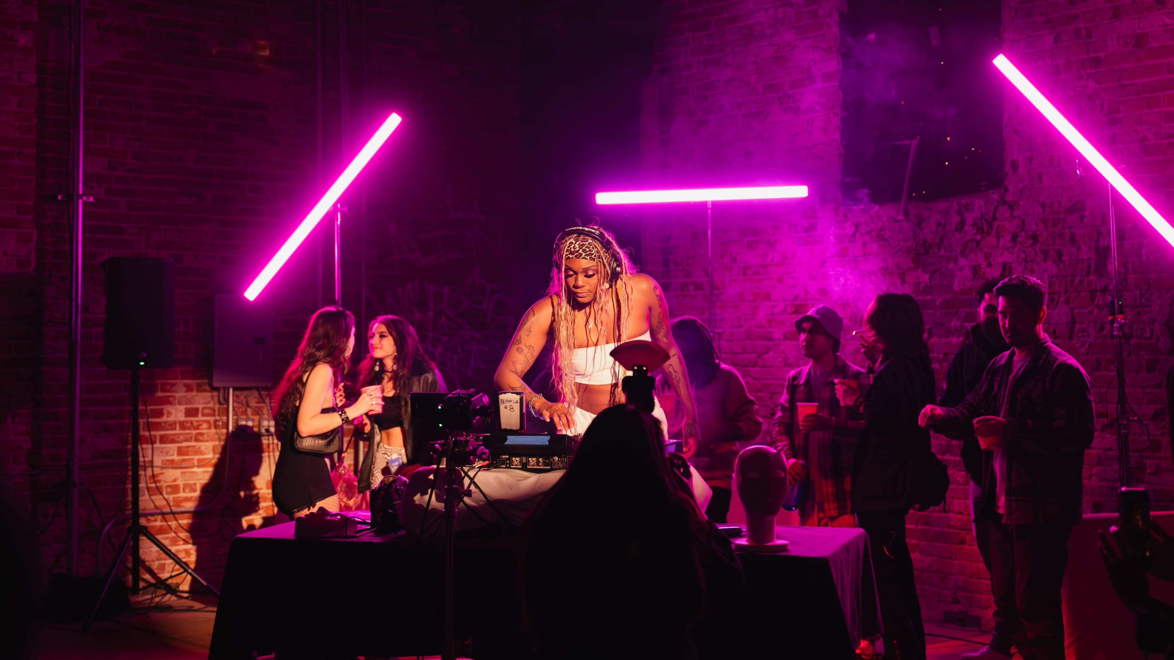 A DJ performs in a dimly lit venue, surrounded by an audience and illuminated by pink neon lights.