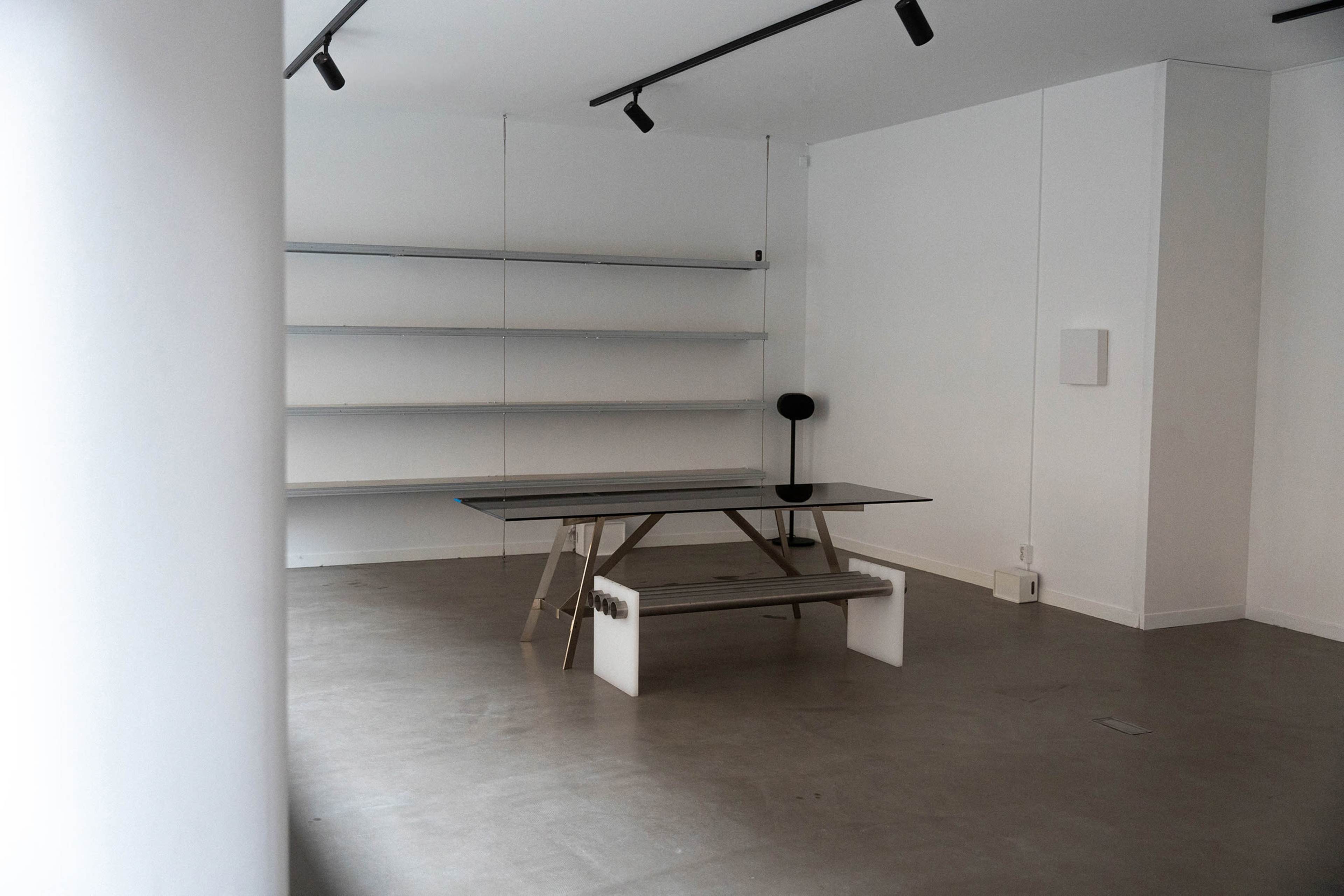 The image shows a minimalist interior of a room with a glass table and a bench, along with white walls and shelves.