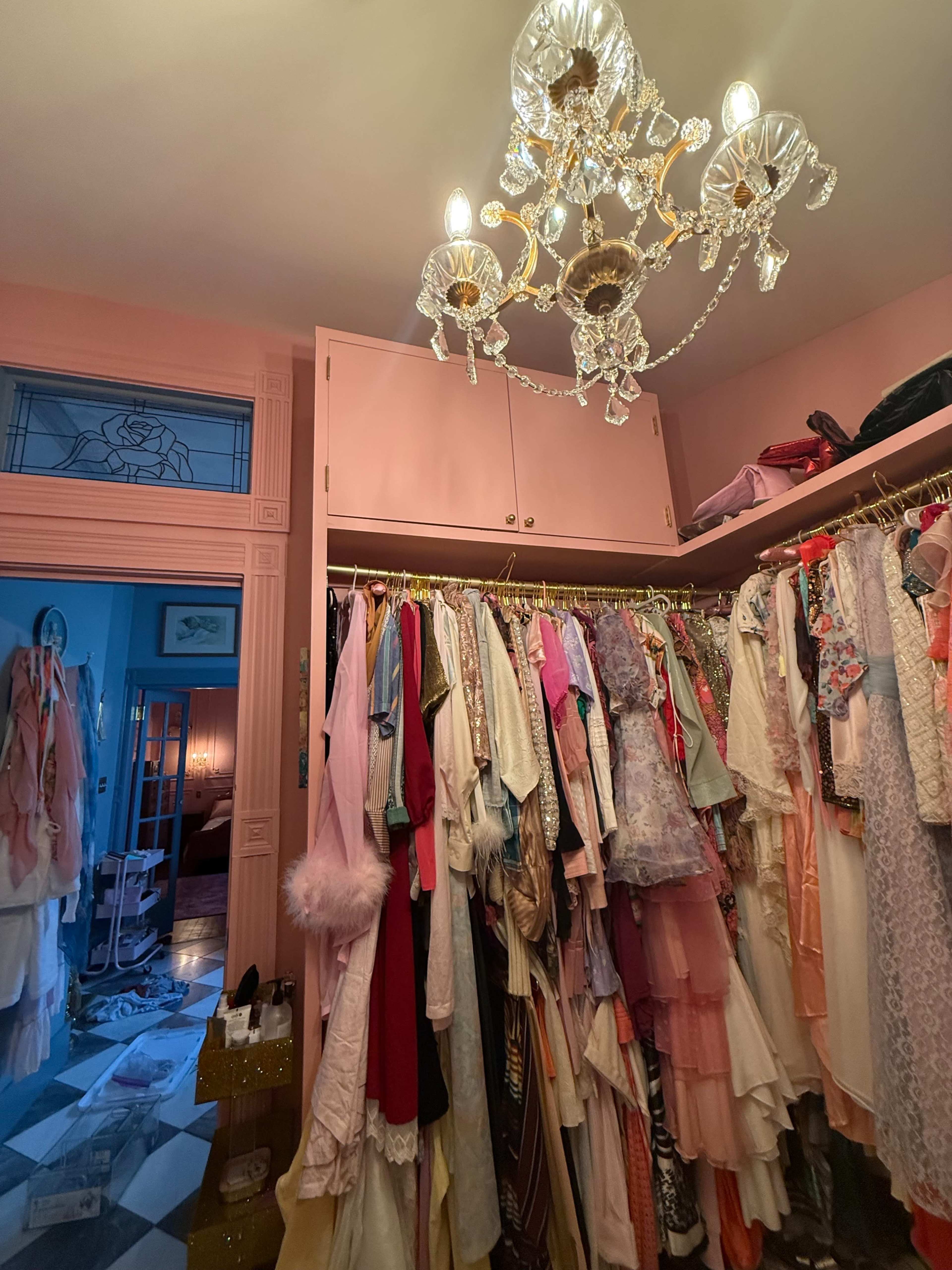 The image shows a closet filled with a variety of colorful clothing hanging on hangers, illuminated by a crystal chandelier above.