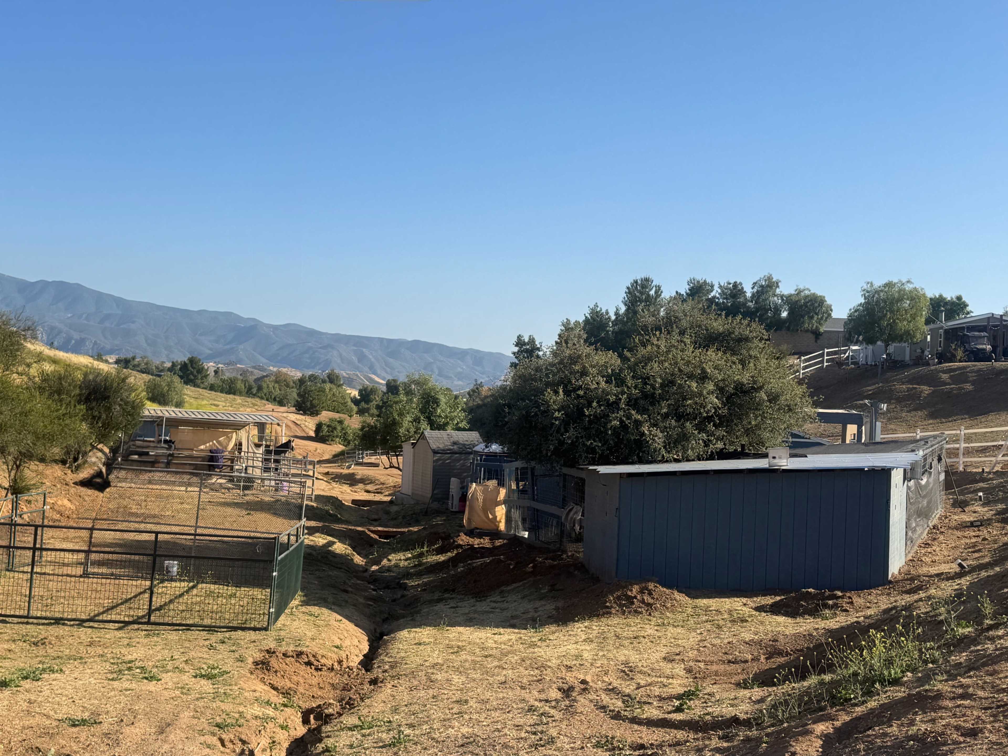 The image shows a landscape with several structures and fenced areas, set against a backdrop of mountains under a clear blue sky.