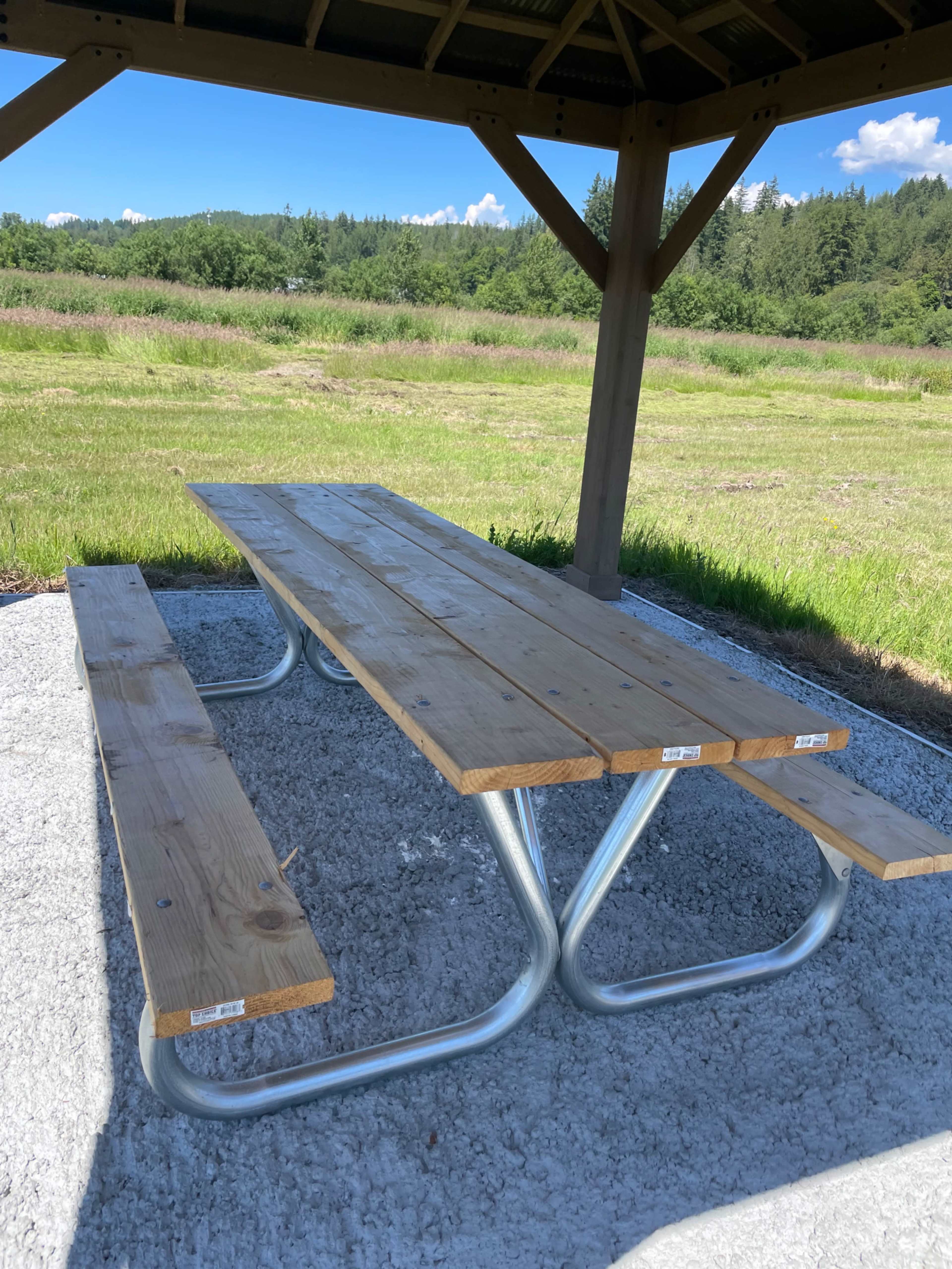 A wooden picnic table with metal supports is situated under a gazebo in a grassy field.