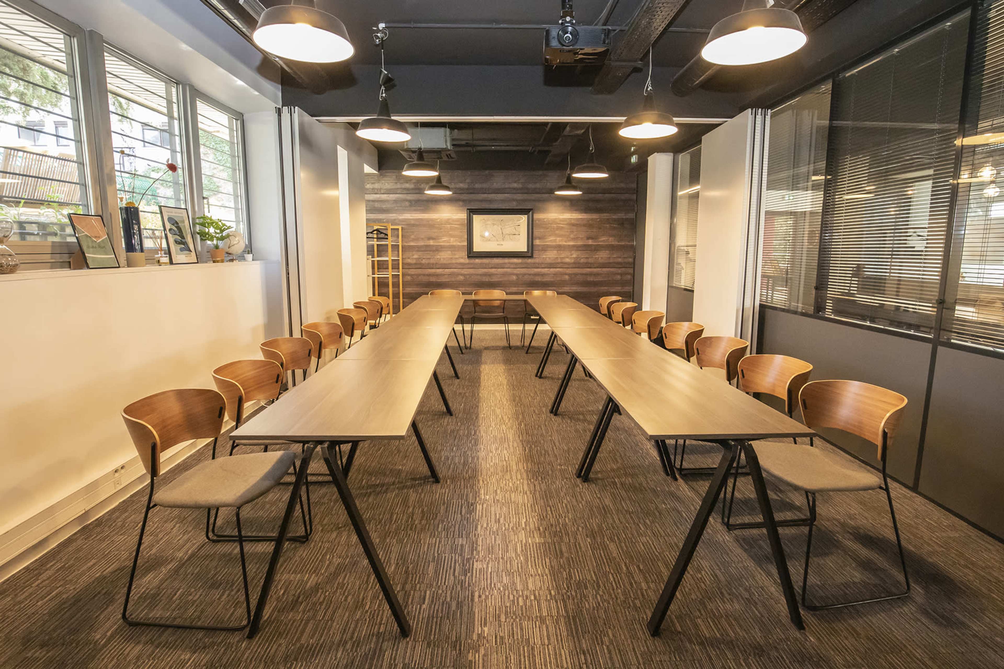 A conference room is set up with two rows of tables and chairs facing each other, surrounded by large windows and diverse wall textures.