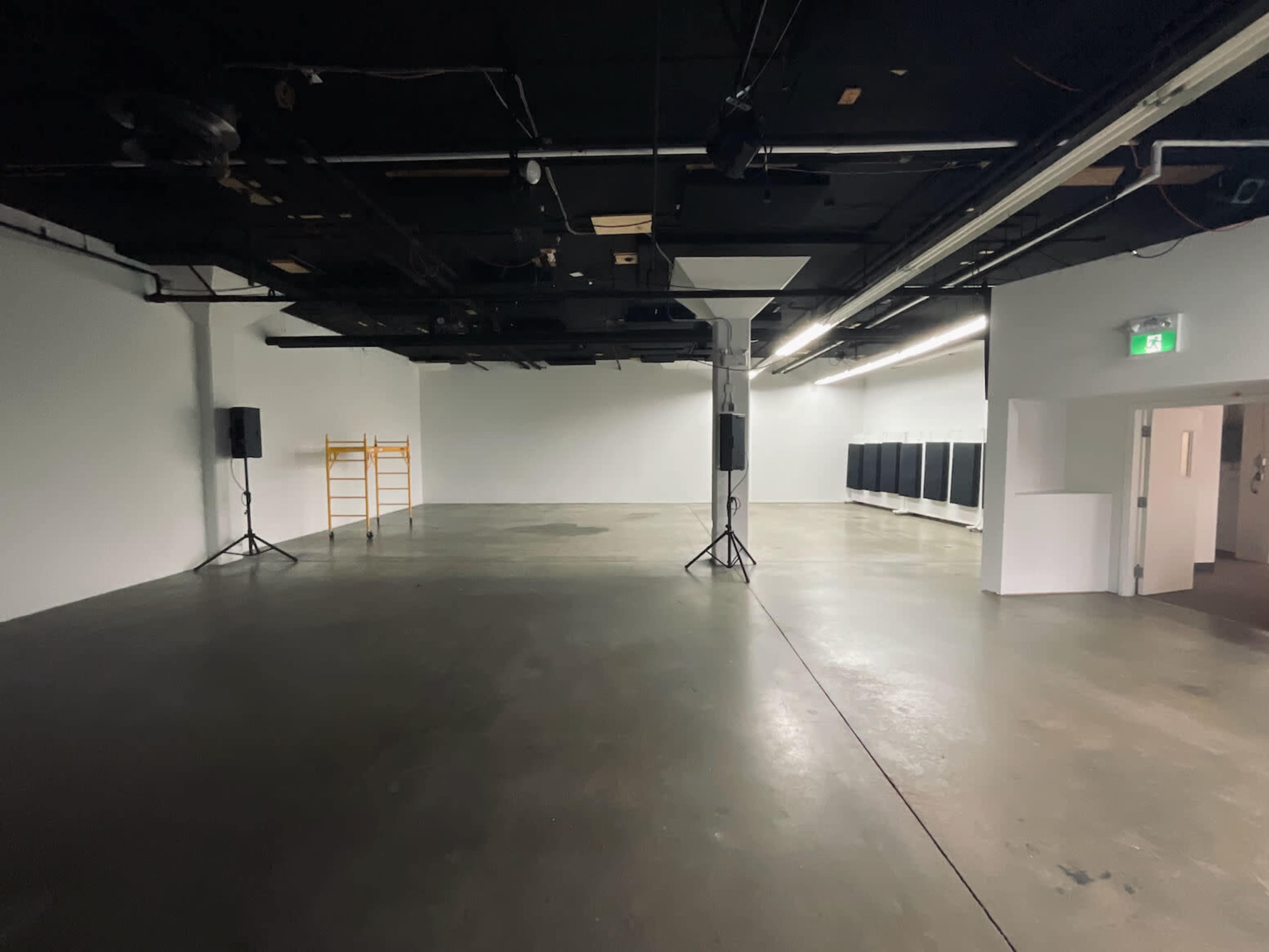 The image shows a large, empty, industrial-style room with concrete floors, white walls, and minimal furnishings, including speakers and a folding platform in one corner.