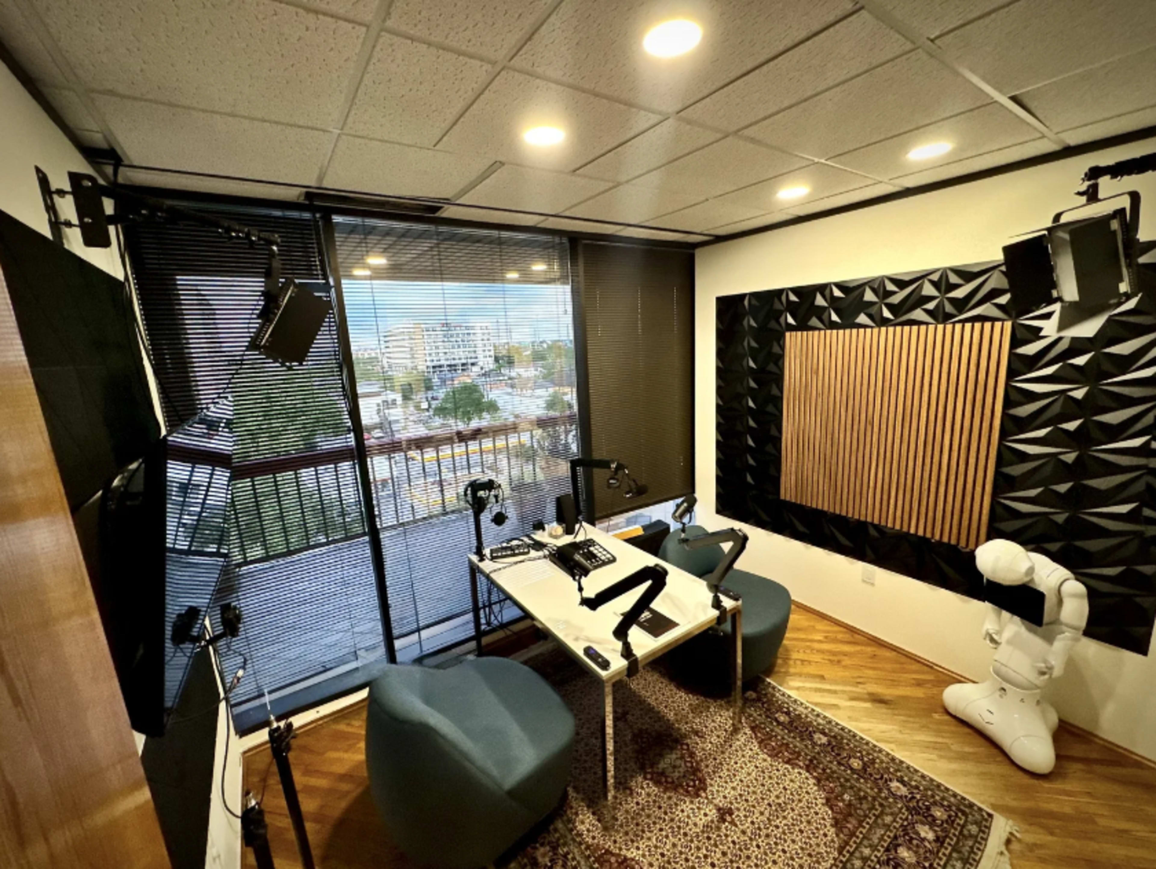 The image shows a recording studio with soundproofing panels, a table equipped with microphones, and a robotic arm, along with a view of the outside through large windows.