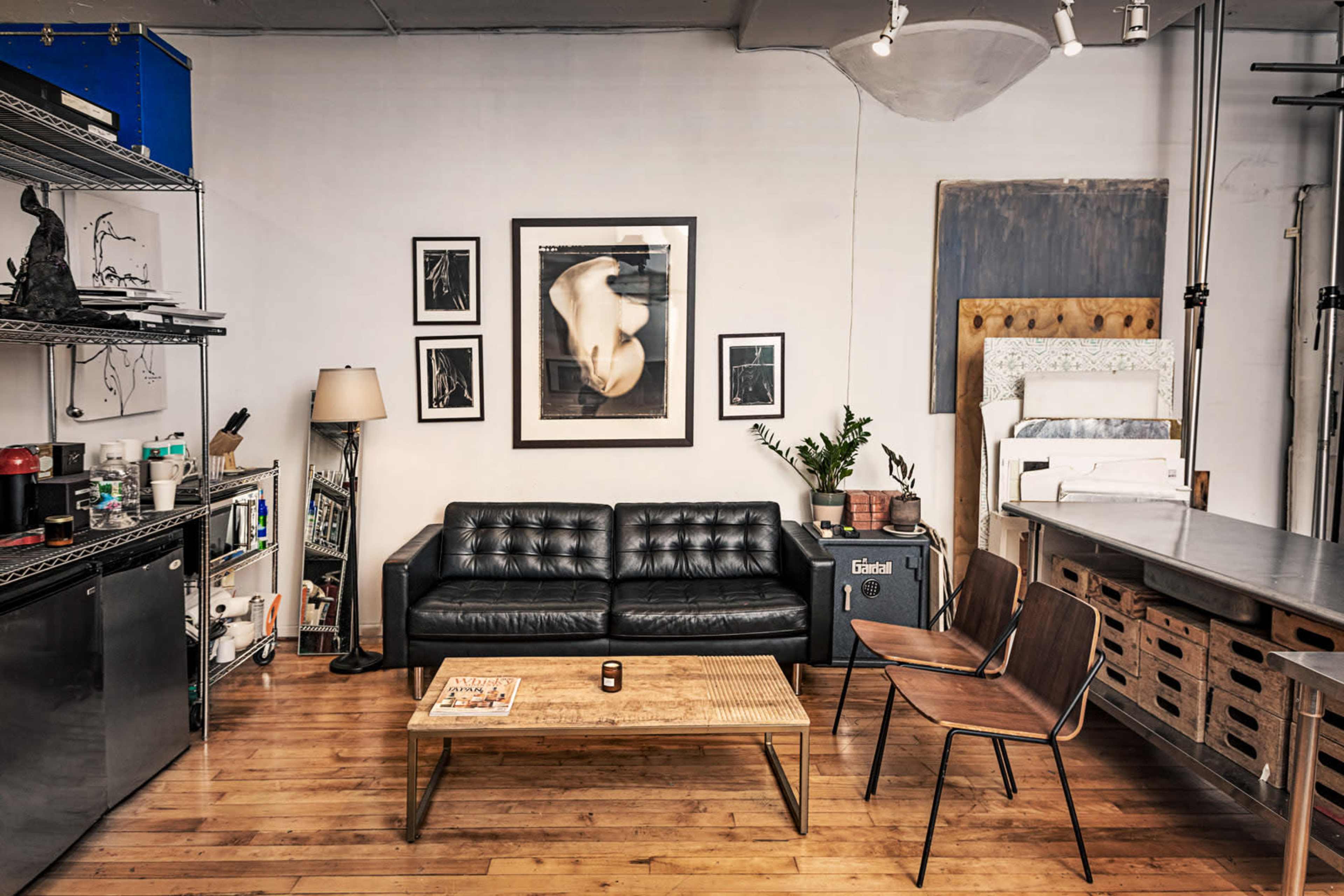 The image shows a modern, industrial-styled living space featuring a black leather sofa, a wooden coffee table, and two wooden chairs, with various artworks and shelving in the background.