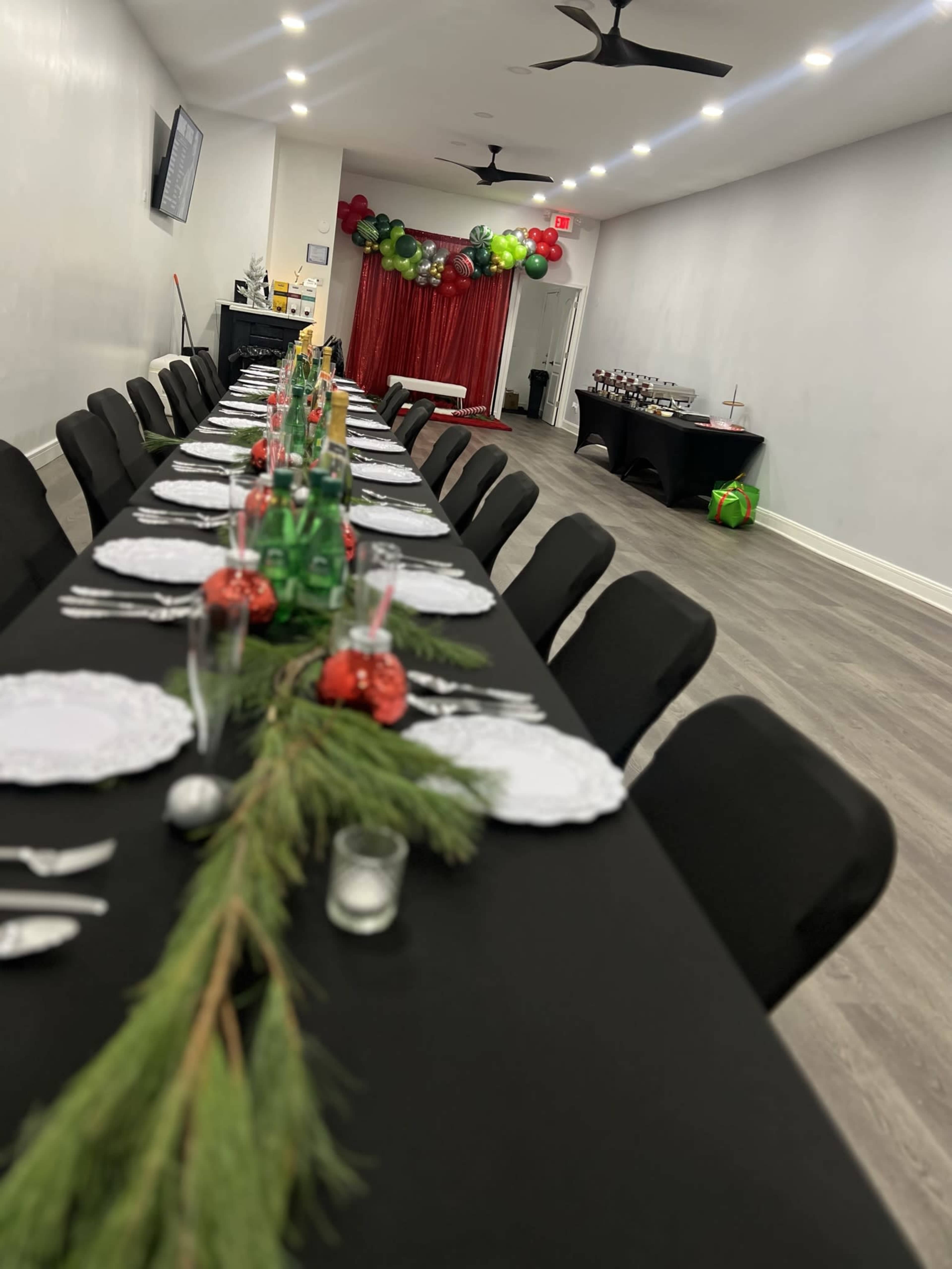 A long table is set for a festive event with green and red decorations, featuring plates, glasses, and a centerpiece of greenery.