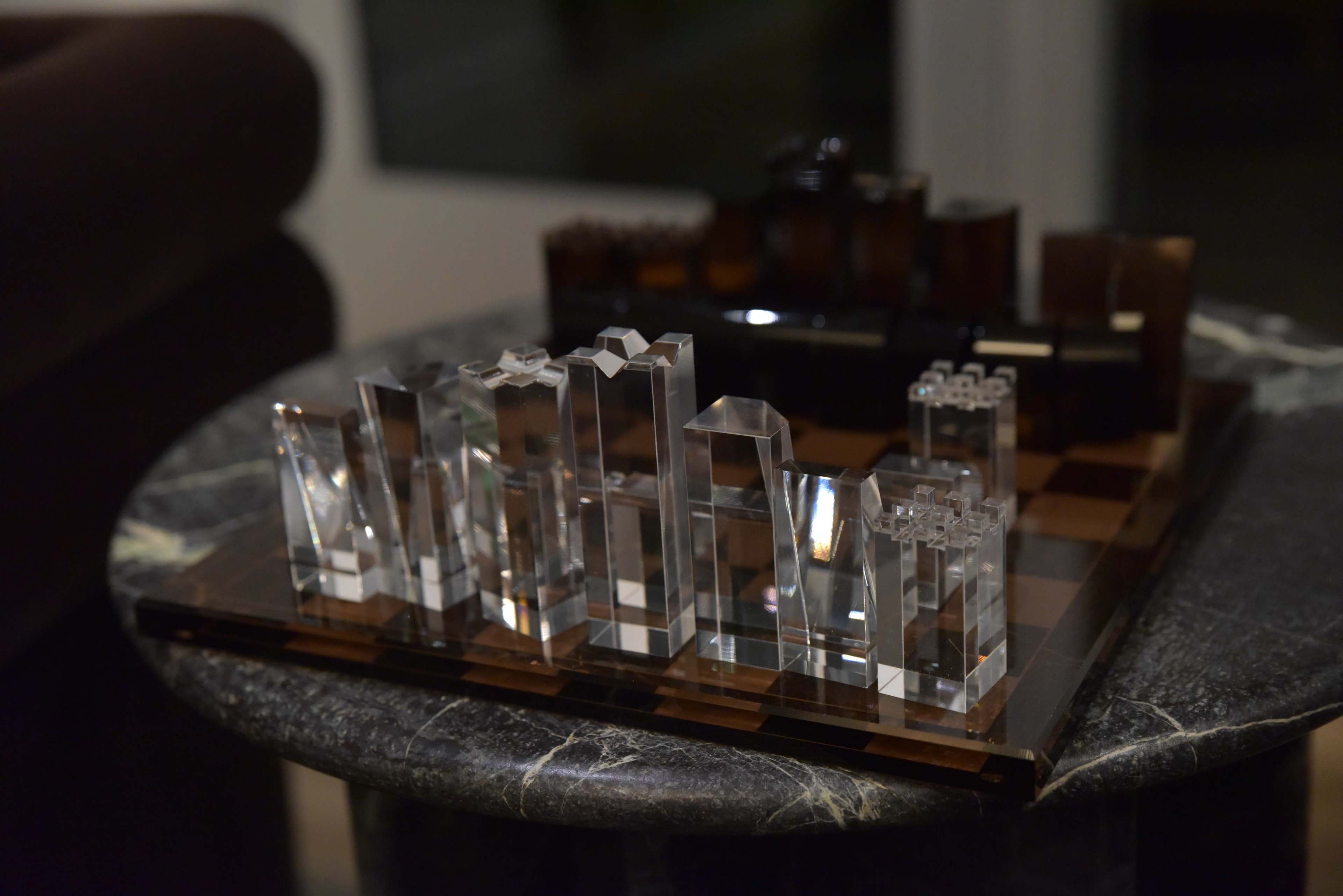 A chessboard with a modern design features clear crystal pieces arranged on a dark marble table.