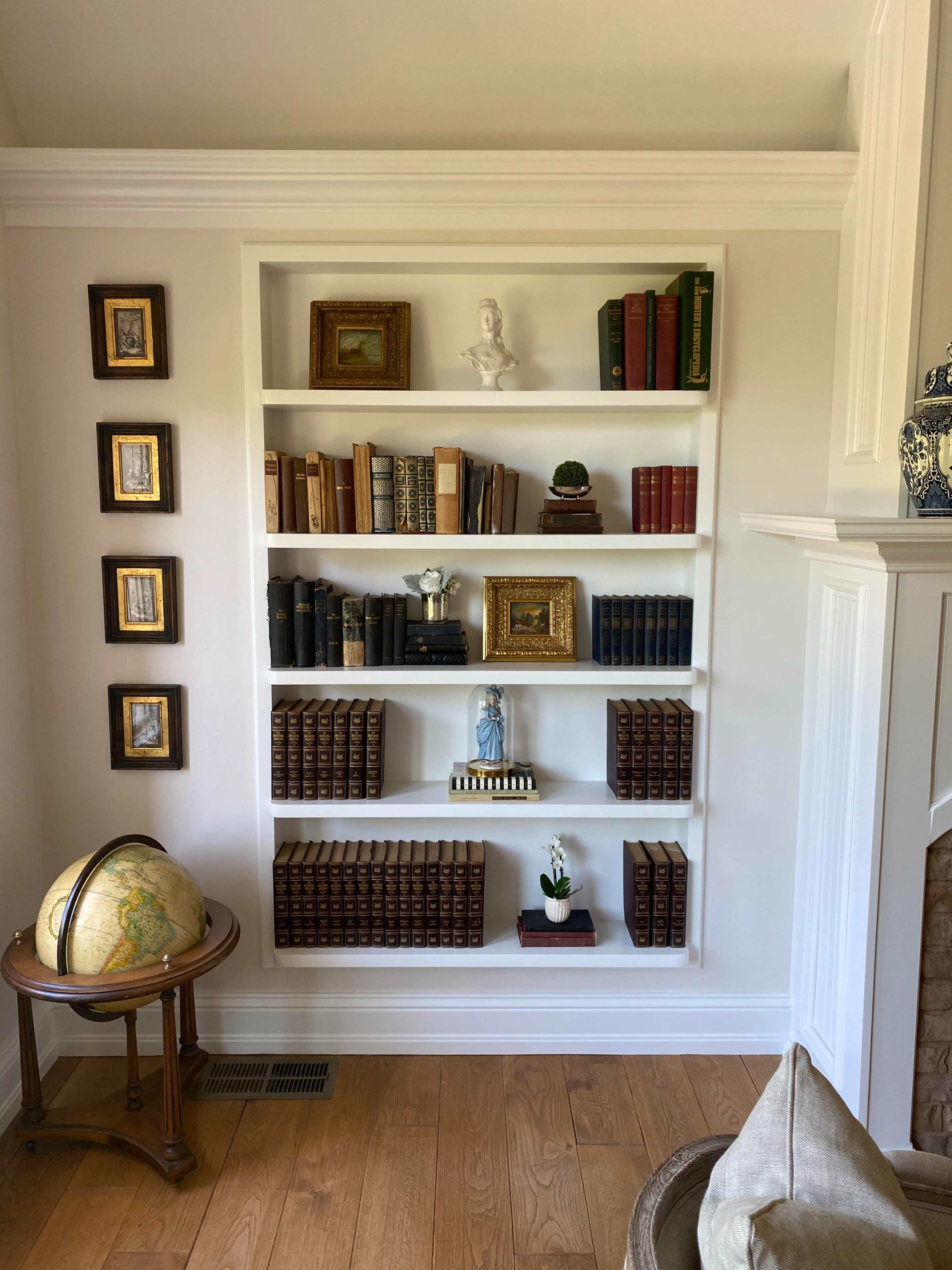 A white bookshelf is filled with various books, decorative objects, and framed art, accompanied by a globe and a small potted plant on a nearby table.