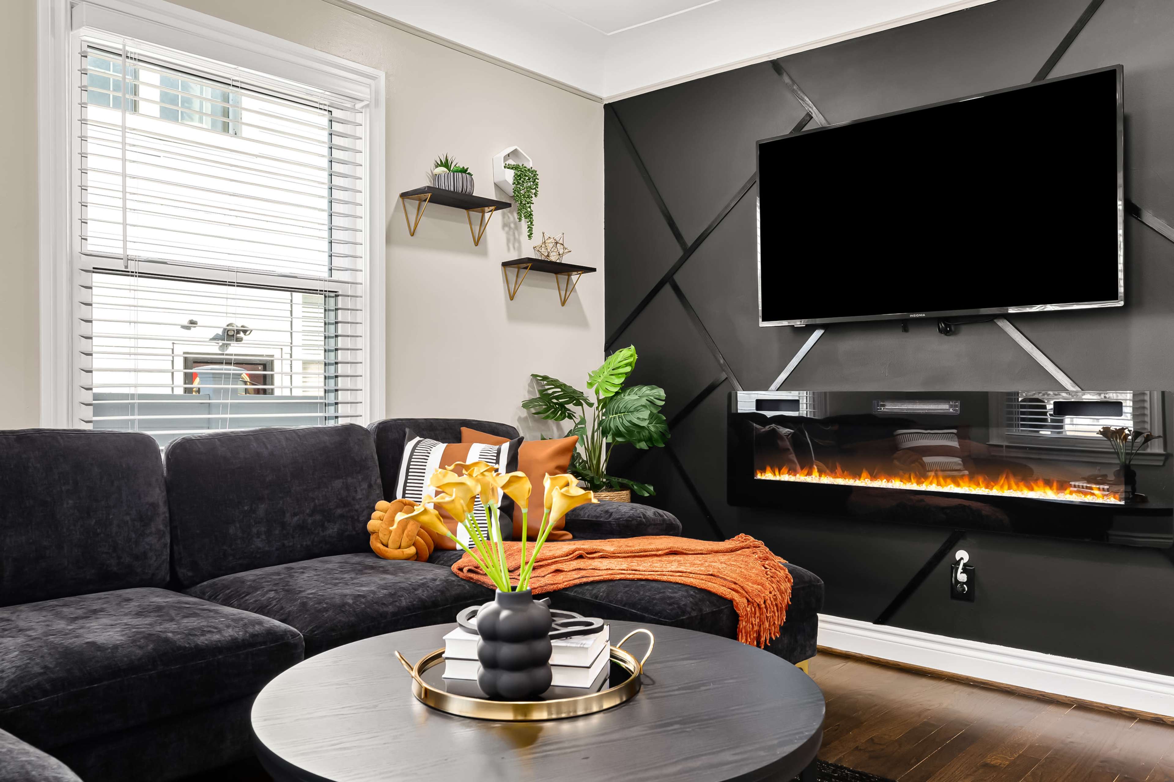 The living room features a dark sectional sofa, a round coffee table, and a mounted TV above an electric fireplace, with decorative plants and shelves on the walls.