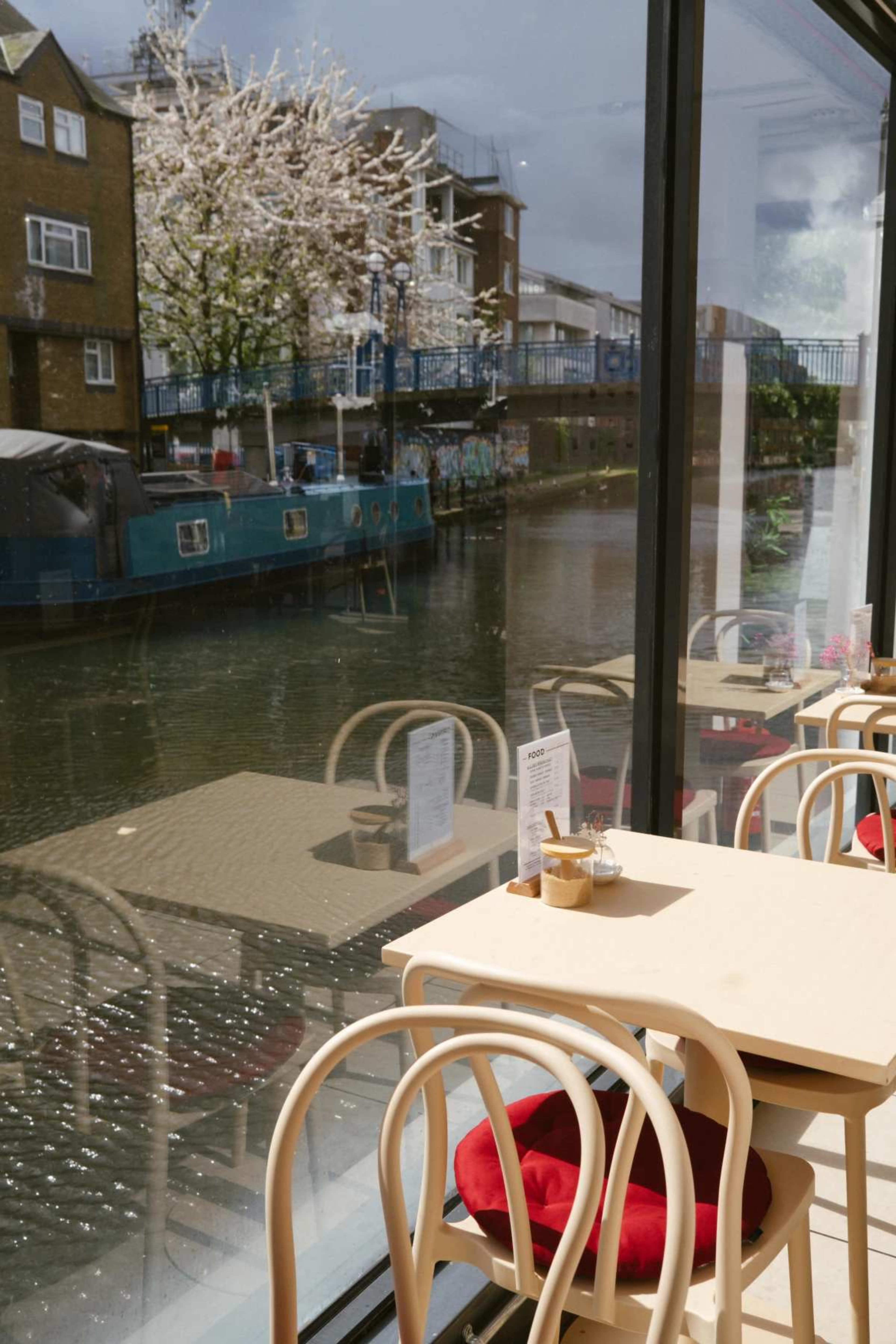 A cafe features wooden tables and red cushions, overlooking a canal with a boat and trees with blossoms.