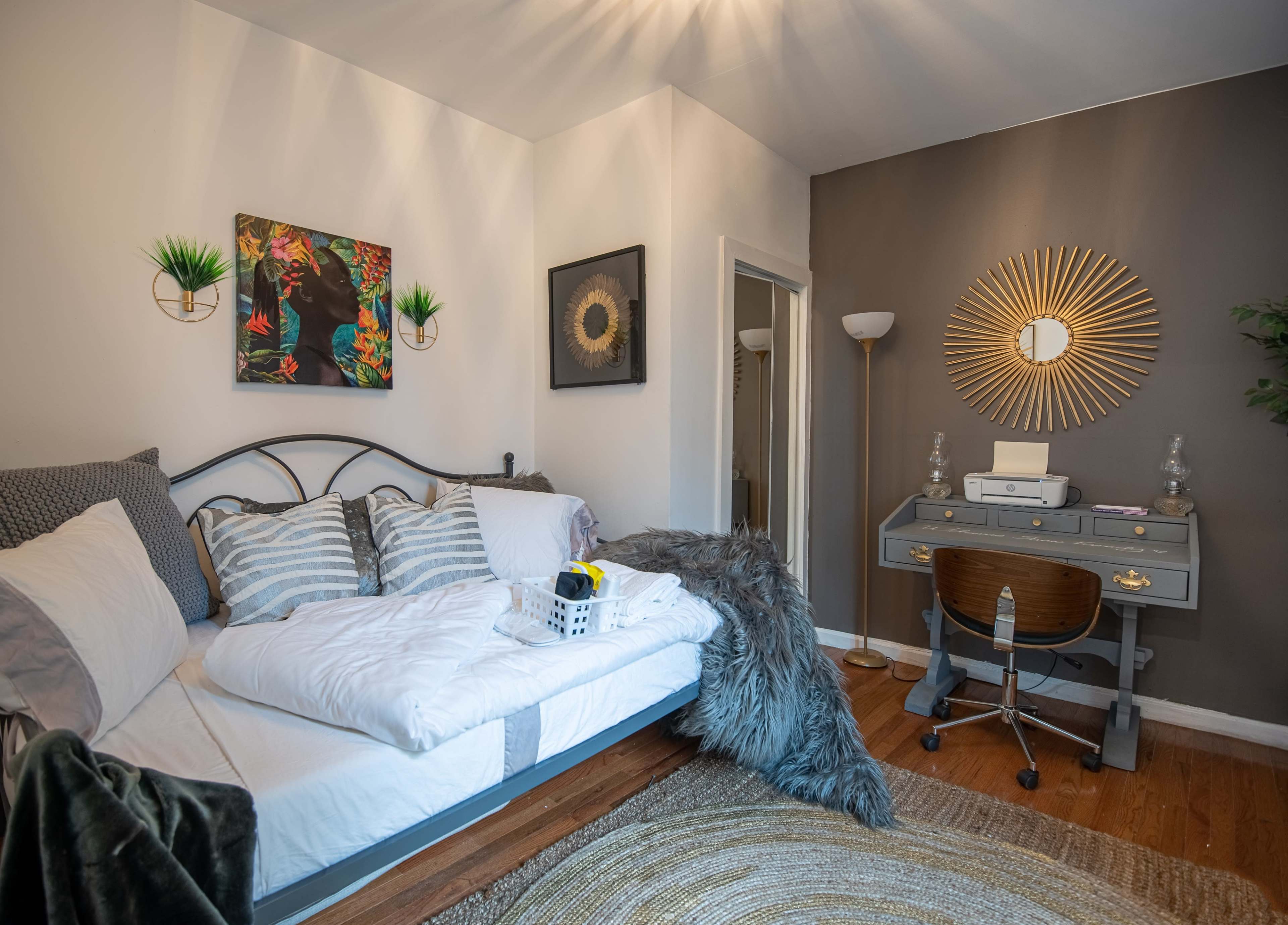 The image shows a cozy bedroom with a bed adorned in white linens, a work desk beside it, and decorative elements on the walls.