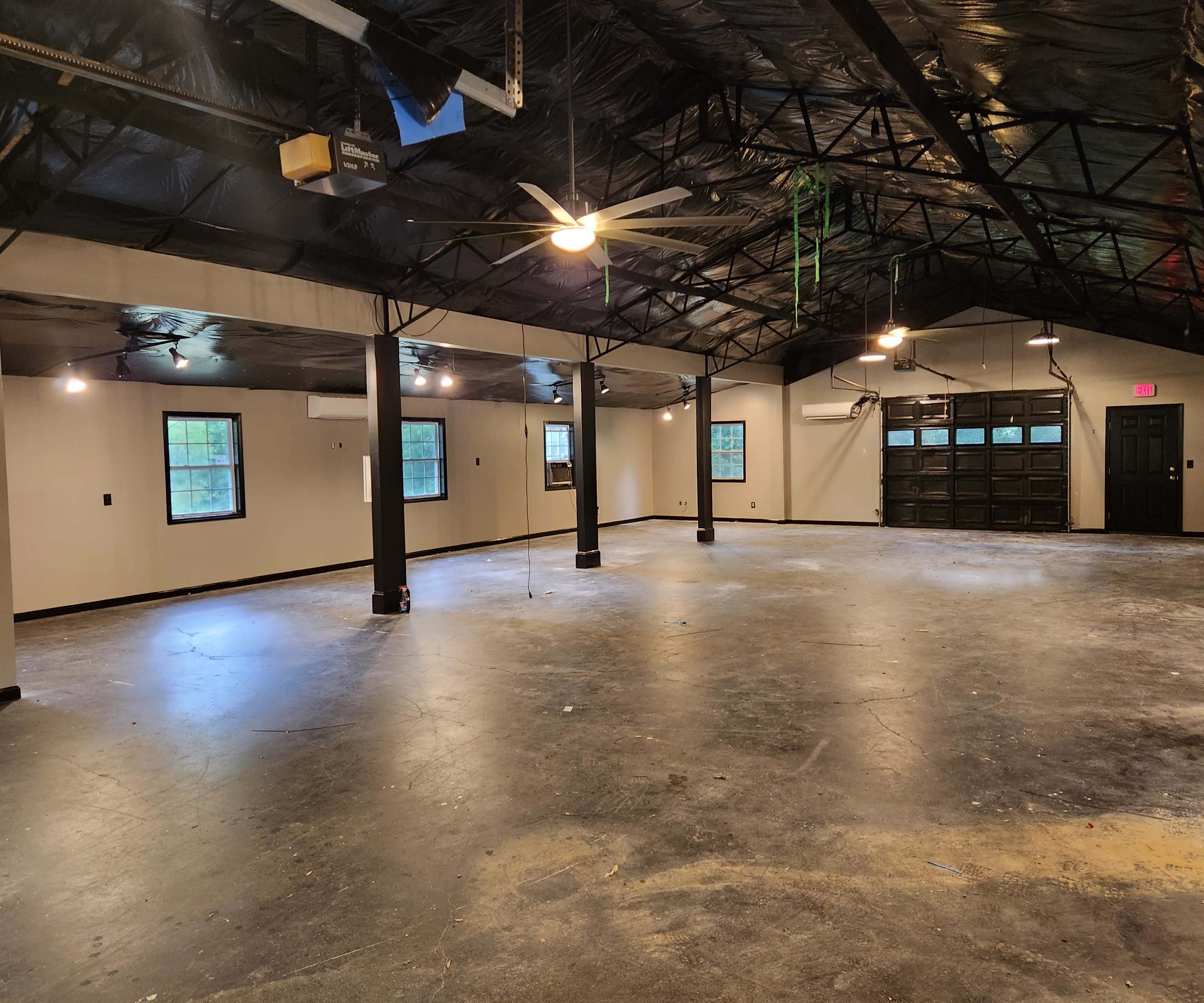 A large, empty room with a high ceiling, exposed beams, and concrete flooring, illuminated by overhead lights and windows.