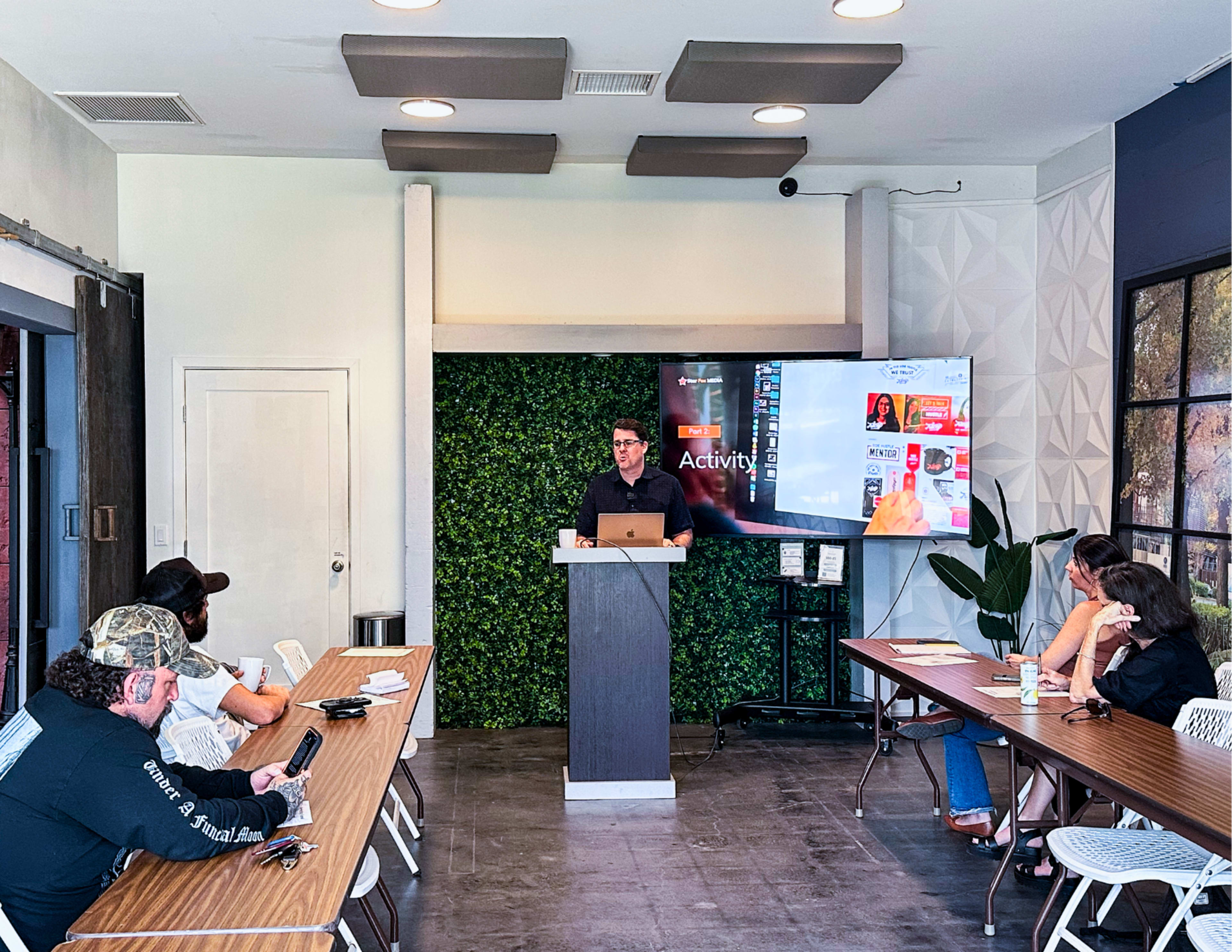 A presenter stands in front of a screen displaying a slideshow while several people sit at a table, observing and taking notes in a meeting room.
